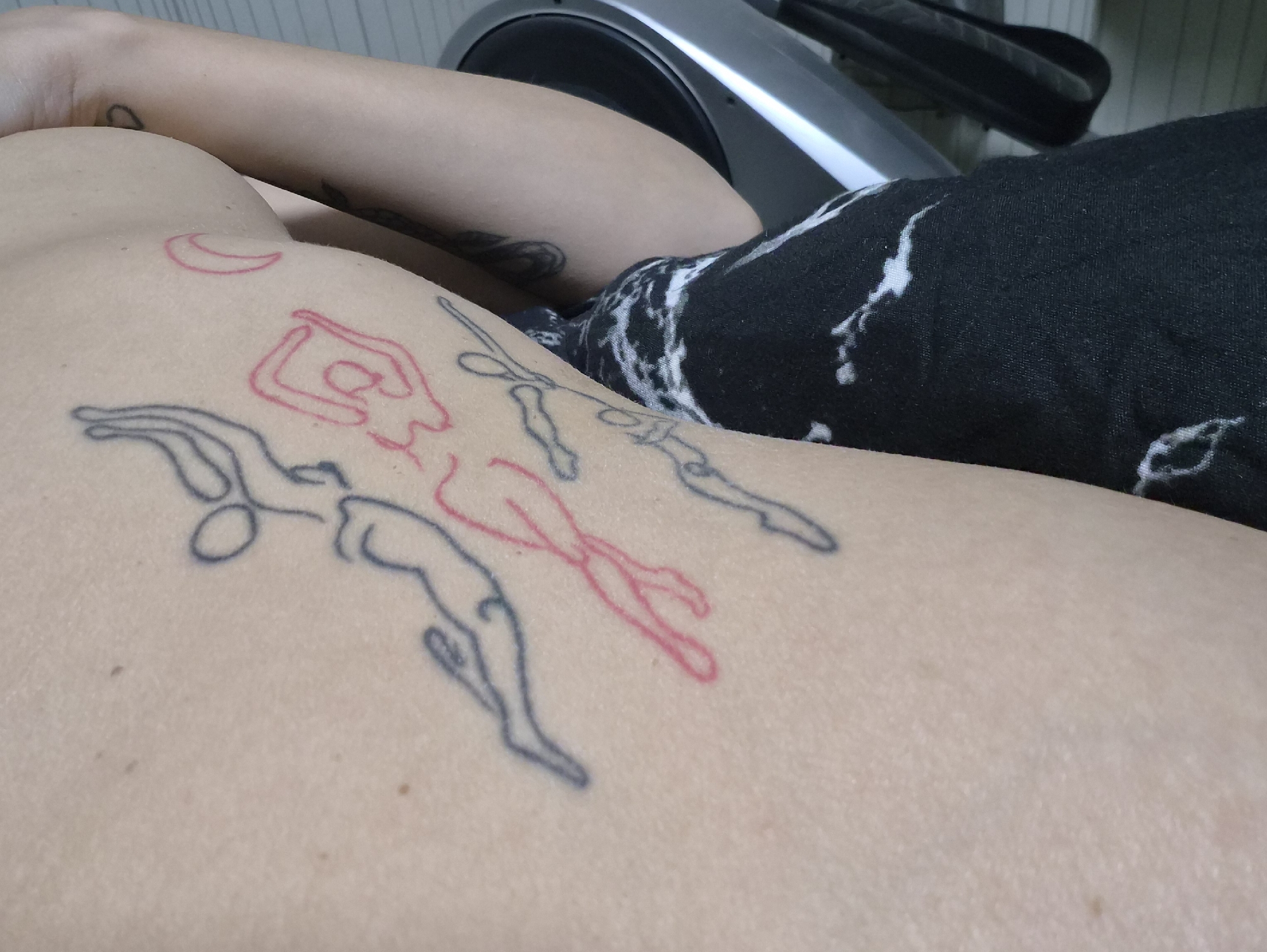 This close-up indoor shot displays a person's upper torso and arm. The skin is light-toned and adorned with several tattoos. On the shoulder/upper back area, there is a red outline tattoo of a crescent moon. Below it, a line of three stylized figures in motion are tattooed; two are outlined in blue/black ink, and one in red ink. Fainter, dark tattoos are also visible on the upper arm closer to the shoulder joint. A piece of black fabric with white, abstract patterns covers a portion of the body, likely clothing. In the background, part of a silver and black exercise machine, possibly an elliptical or bicycle, is visible, suggesting an indoor setting such as a home or gym. The person appears to be resting or lying down. No specific activity is taking place. The lighting is consistent with an indoor environment. No text is visible in the image. The location context for this media file is Chaikovsky, Russia.