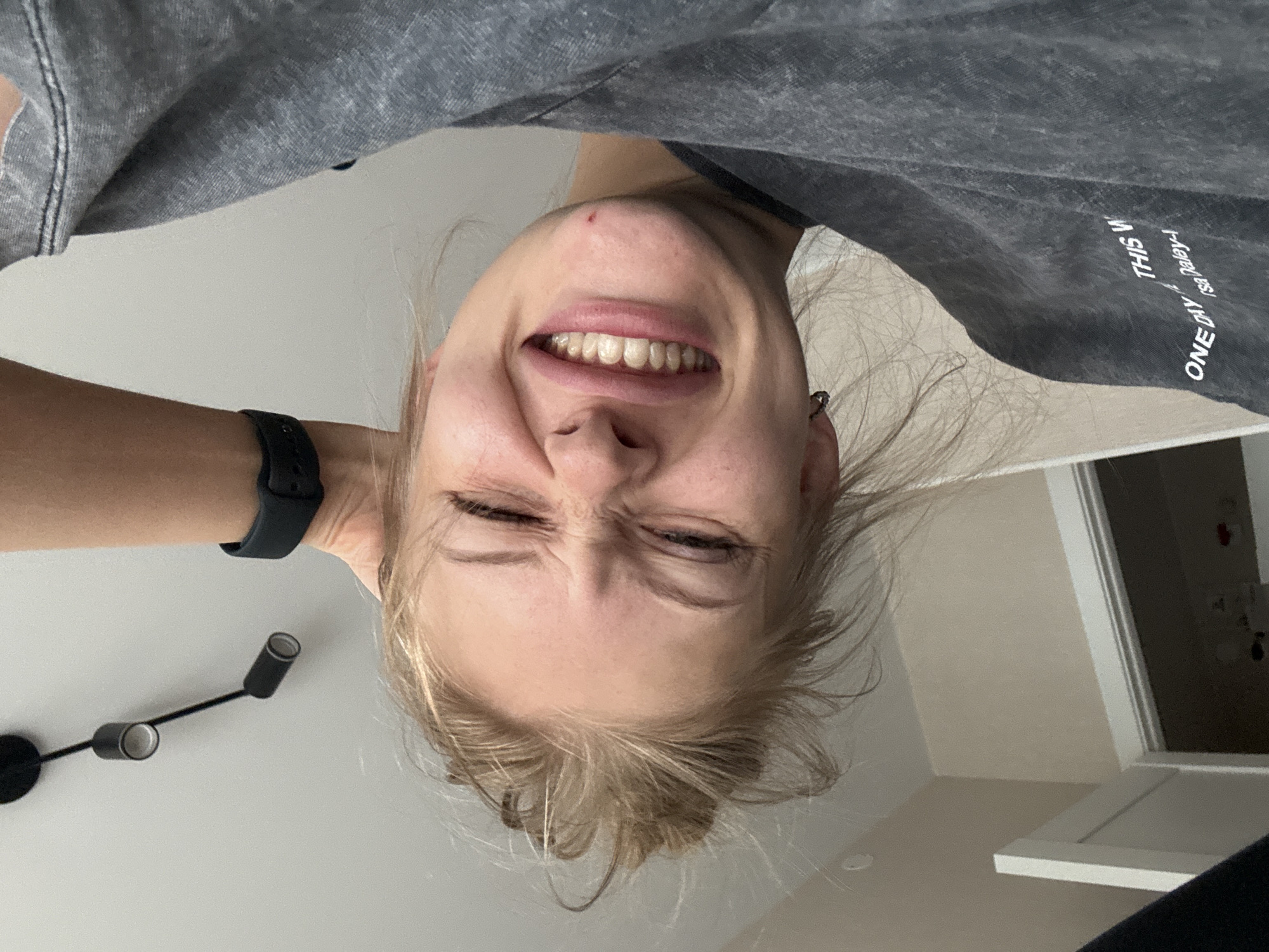 The image shows a young woman with blonde hair tied up in a bun, winking and smiling broadly, with her head tilted to the side. She is wearing a dark grey t-shirt and a black fitness tracker on her left wrist. The background includes a white ceiling with black track lighting, a light-colored wall, and a glimpse of a doorway with a white frame. The image appears to be a selfie taken indoors, likely in a home or apartment. There is no indication of the city of Perm, Russia, or any specific urban environment in the image.