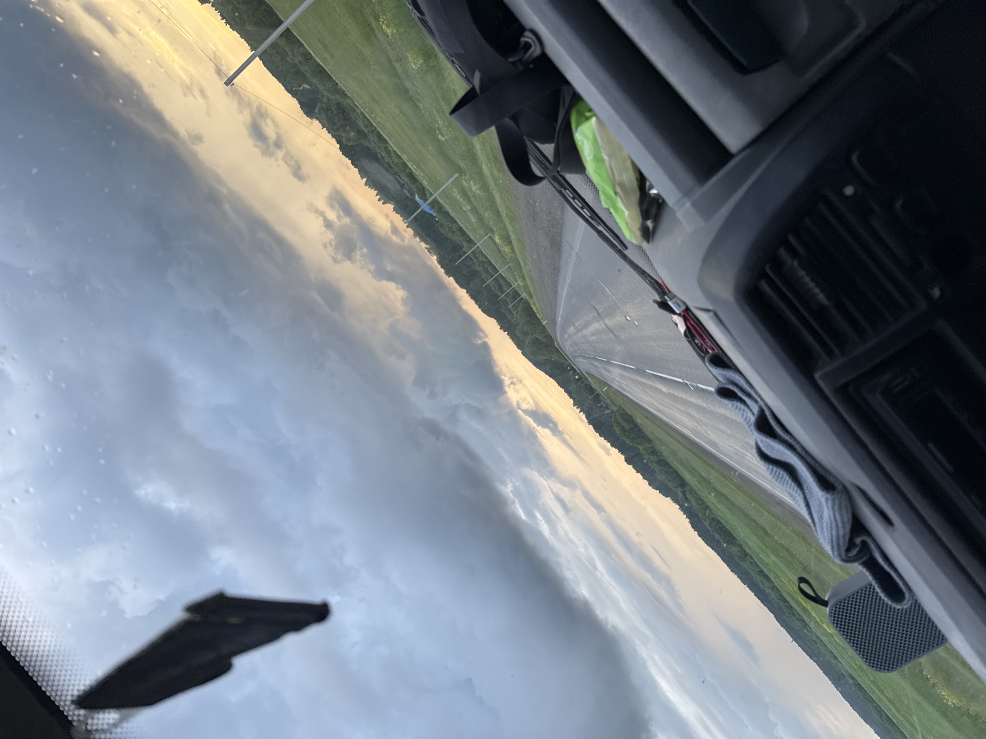 The image depicts a view from inside a vehicle, looking out through the windshield. The perspective is angled, with the right side of the frame appearing closer and the left side receding.
The foreground shows the dashboard of the vehicle, with a gray textile folded neatly on top. To the left of the textile is a small, dark gray, mesh-textured speaker. Further to the left, hanging from the top of the windshield, is a dark, irregularly shaped object, possibly an air freshener.
The middle ground is dominated by a road stretching into the distance, with white lane markings visible. The road surface appears wet, suggesting recent rain or damp conditions. To either side of the road are grassy fields, which appear lush and green. A line of trees forms a backdrop to the fields on both sides, receding into the distance.
The background features a dramatic sky. On the left side, dark, heavy clouds dominate, suggesting an approaching storm or lingering rain. These clouds transition to lighter grays and whites towards the center. On the right side of the sky, a warm, golden light breaks through, indicative of sunset or sunrise. This light illuminates the tops of the clouds and casts a glow over the horizon. Several utility poles with wires are visible along the right side of the road, extending into the distance.
The overall impression is one of driving through a landscape with changing weather conditions, likely during the evening or early morning. The mention of Chernovskoe, Russia, provides a geographical context, suggesting this scene is from that specific location.