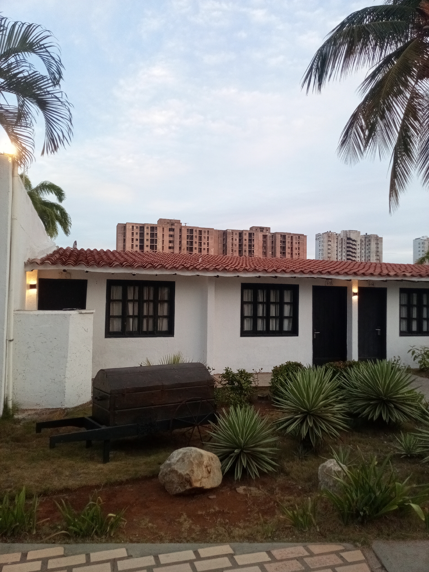 This image presents a serene outdoor scene, likely captured in Porlamar, Venezuela. In the foreground, a single-story white building with a distinctive red tiled roof stands prominently. The building features several dark-framed windows with light-colored curtains and two dark doors, above which outdoor sconce lights are illuminated. Visible above the doors are the numbers "1-15" and "1-14".
A manicured garden occupies the space in front of the building, featuring several large, spiky succulent plants, possibly Agave or Yucca, alongside smaller green bushes and patches of grass on reddish-brown soil. A few natural rocks are scattered within the garden. A notable object in the garden is a dark, antique-looking wooden chest, resting on a black metal cart or wheelbarrow, positioned to the left of the building's center.
Palm fronds partially frame the scene from the top left and right, emphasizing the tropical or subtropical setting. In the background, several tall, multi-story apartment or commercial buildings rise, indicating an urban or semi-urban environment beyond the immediate garden and low-rise structure.
The sky is a soft, light blue with scattered white clouds, and the diffused lighting suggests either early morning or late afternoon/dusk, with the artificial lights on the building subtly glowing. No people or specific activities are visible, contributing to a tranquil and still atmosphere.