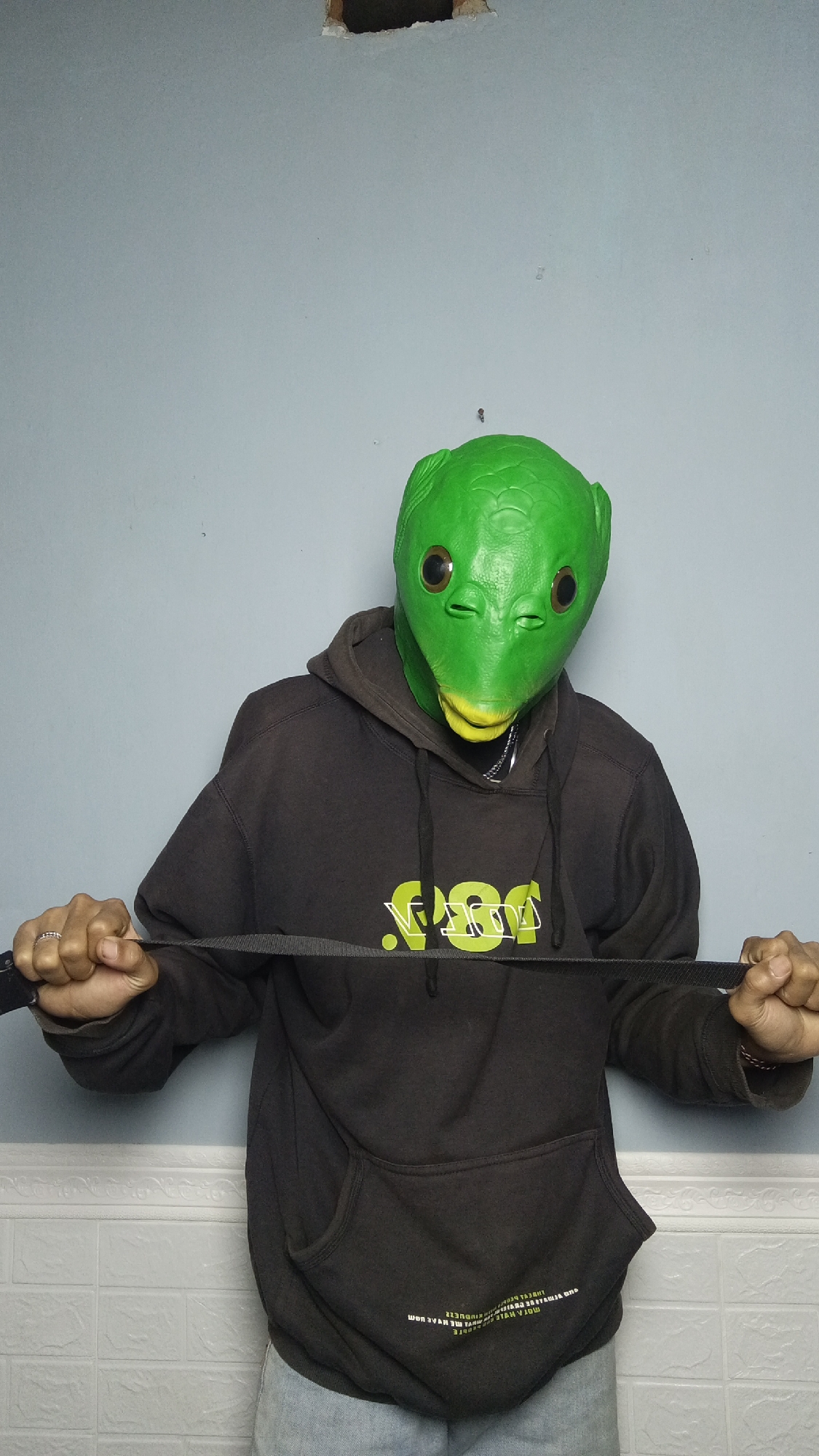 A person wearing a green alien mask and a dark hooded sweatshirt stands in front of a light blue wall. The person is holding a black strap with both hands, which is stretched horizontally across their chest. The background wall has a visible hole near the top. The lower part of the image shows a tiled wall with a white brick pattern. The scene appears to be an indoor setting, possibly a room or studio, located in Kebumen, Indonesia.