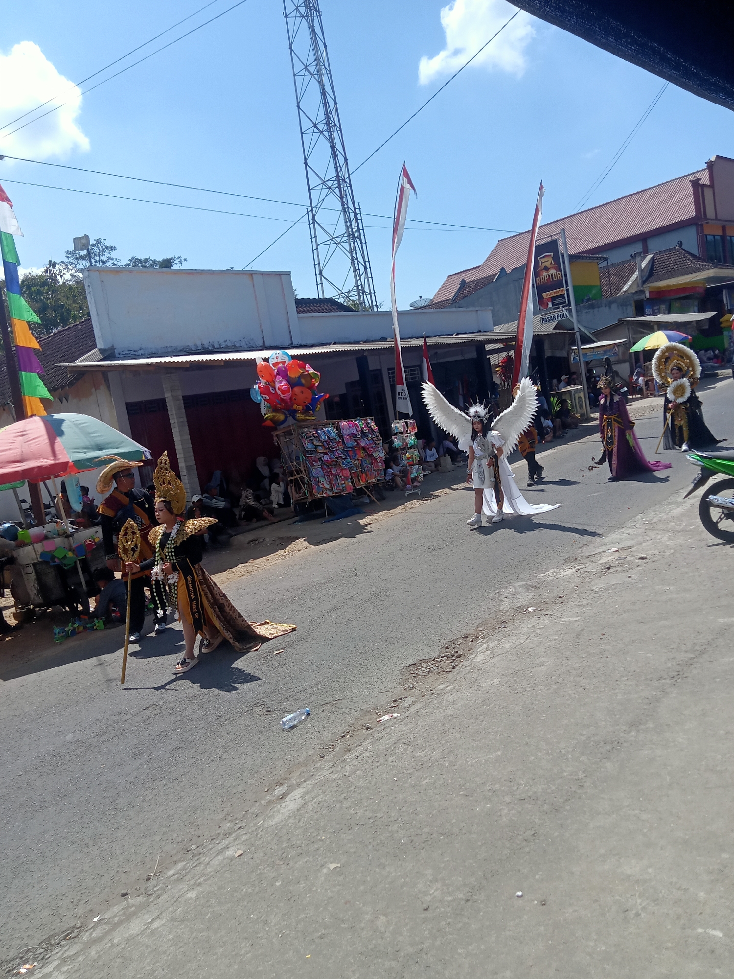 In Pule, Indonesia, on a sunny day, a street parade is in progress. People are dressed in elaborate costumes, some with large wings, reminiscent of angels or mythical figures. Others are adorned in traditional Indonesian attire, complete with ornate headdresses. They walk along a paved road, with spectators lining the sides. In the background, buildings with tiled roofs and a tall radio tower are visible. Colorful flags and balloons add to the festive atmosphere. Stalls selling various goods, including balloons and toys, are set up along the roadside. A green motorcycle is parked on the right side of the road. The scene captures a vibrant cultural event taking place in a seemingly rural or semi-urban setting in Indonesia.