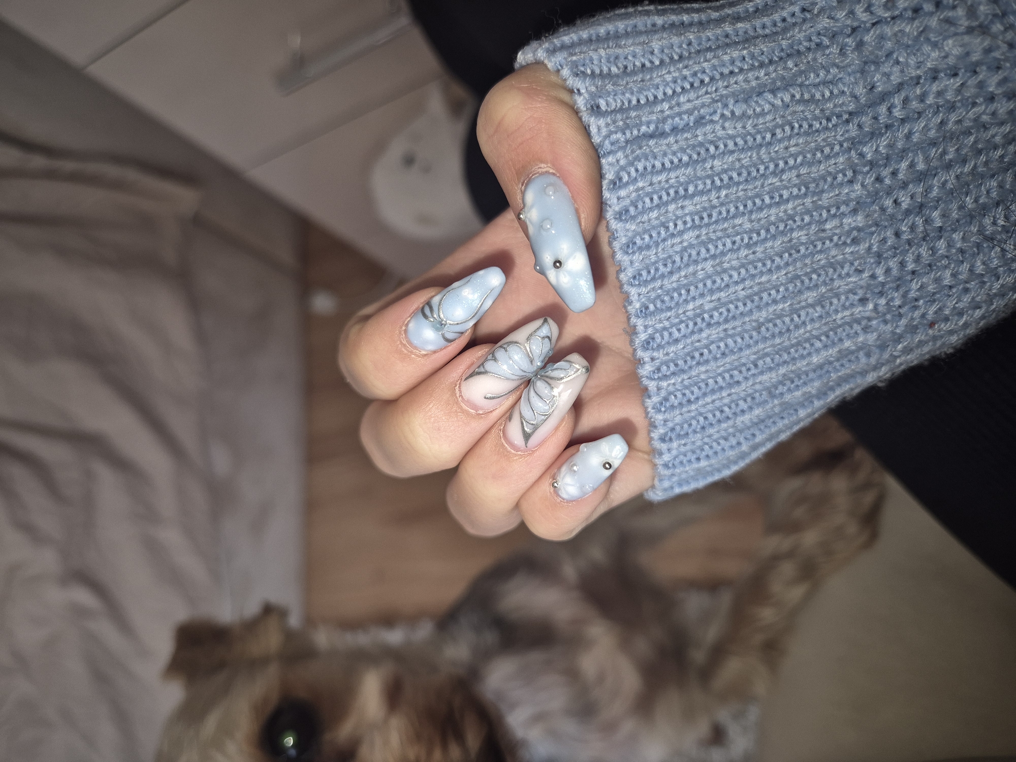 A hand wearing a light blue knit sweater is holding out fingers with light blue and white manicured nails. The nail art includes butterfly designs and small silver studs. The background includes a blurry image of a light brown dog, a cream-colored bed, and a white object. The location is not specified.