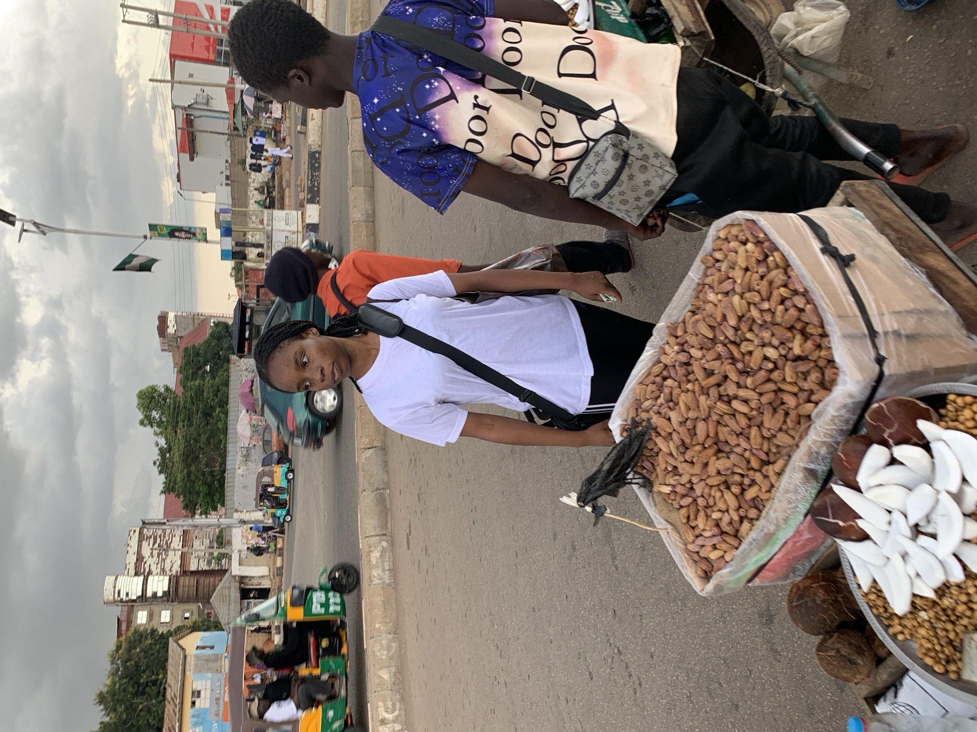 In Jos, Nigeria, a busy street scene unfolds under a cloudy sky. In the foreground, a young woman with braided hair, wearing a white t-shirt and black pants, stands facing left. She has a black crossbody bag slung over her shoulder. Beside her, a vendor's cart displays a large transparent bag overflowing with dates, and a metal tray filled with nuts and sliced coconut. A dead bird, perched on a stick, is positioned atop the dates, its wings spread. Behind the woman, a young man in a blue t-shirt with a white and pink pattern featuring repeating "Door" text and a grey patterned crossbody bag stands facing away from the camera. Another person in an orange shirt is partially visible behind him. The background reveals a typical urban environment with paved roads, sidewalks, and buildings, including a distinct white cylindrical structure. Several vehicles, including a green and yellow auto-rickshaw and a green car, are visible on the road, suggesting ongoing traffic. The overall impression is one of everyday commerce and activity in a Nigerian city.