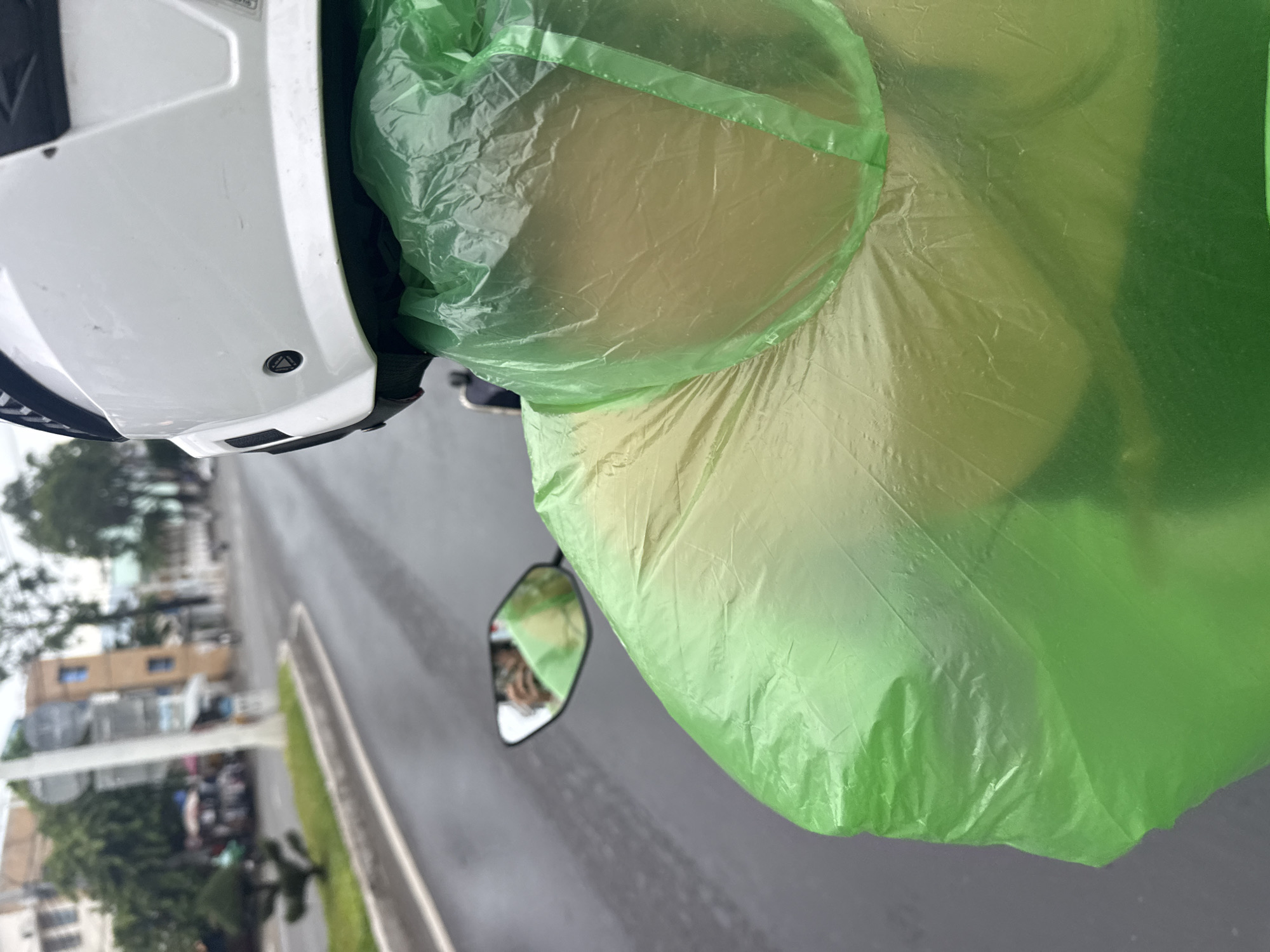 The image shows a close-up, first-person perspective of someone riding a motorcycle in Nha Trang, Vietnam. The rider is wearing a white helmet and a bright green, translucent raincoat. The raincoat is bunched up around the rider's head and shoulders. A motorcycle rearview mirror is visible on the left, reflecting the rider's face, also covered by the green raincoat and helmet. The road is wet and appears to have just rained, with tire tracks visible. In the background, buildings and trees line the street, suggesting an urban environment. The scene is captured from a low angle, emphasizing the rider's immediate surroundings.