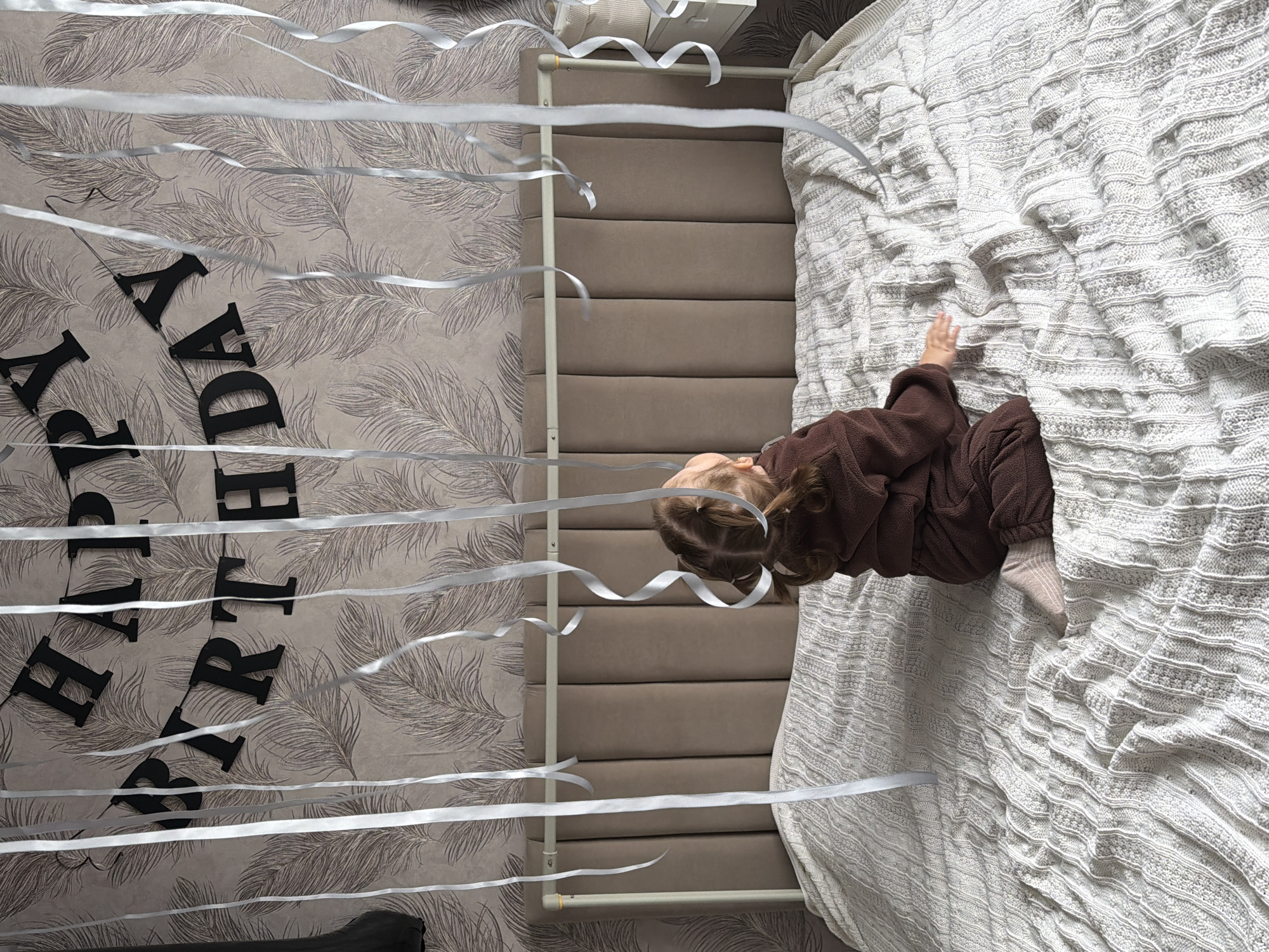 An overhead shot, rotated 90 degrees clockwise, captures a young child lying on a bed in a room decorated for a birthday celebration. The child, likely a toddler or preschooler, is dressed in a brown, textured outfit and has light brown hair styled in pigtails. They are positioned on their side on a large, light-colored, possibly knitted or crocheted, blanket that covers the bed, with one arm slightly raised as if reaching or observing.
Numerous silver ribbons or streamers are strung across the room, creating a festive canopy above the bed and leading towards a wall. This wall is adorned with patterned wallpaper featuring subtle, light-toned feather or leaf designs. Against this wallpaper, black cutout letters spell out "HAPPY BIRTHDAY," though parts of "BIRTH" are obscured by ribbons and the composition, "HAPPY" and "DAY" are clearly visible.
Behind the child's head is a brown, padded headboard, and a white metal frame or railing is visible, possibly part of a bed or crib structure. The lighting suggests an indoor scene, likely during the day. The overall impression is a quiet, celebratory moment, possibly an early morning birthday surprise, in a home in Reshetnikovo, Russia.