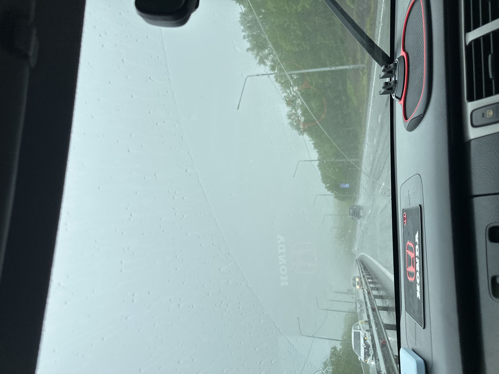 The image captures a view from inside a vehicle driving on a highway in Svetly, Russia, during rainy weather. Water droplets are visible on the windshield, obscuring the view of the road ahead. The highway is wet, and other vehicles are visible in the distance. On the dashboard, a red and black mat with the "HONDA" logo is present. The sky is overcast, contributing to the overall dim lighting. The surrounding environment appears to be a mix of trees and streetlights, indicating an urban or suburban setting. No people are clearly visible within the frame, apart from the implied driver of the vehicle.
