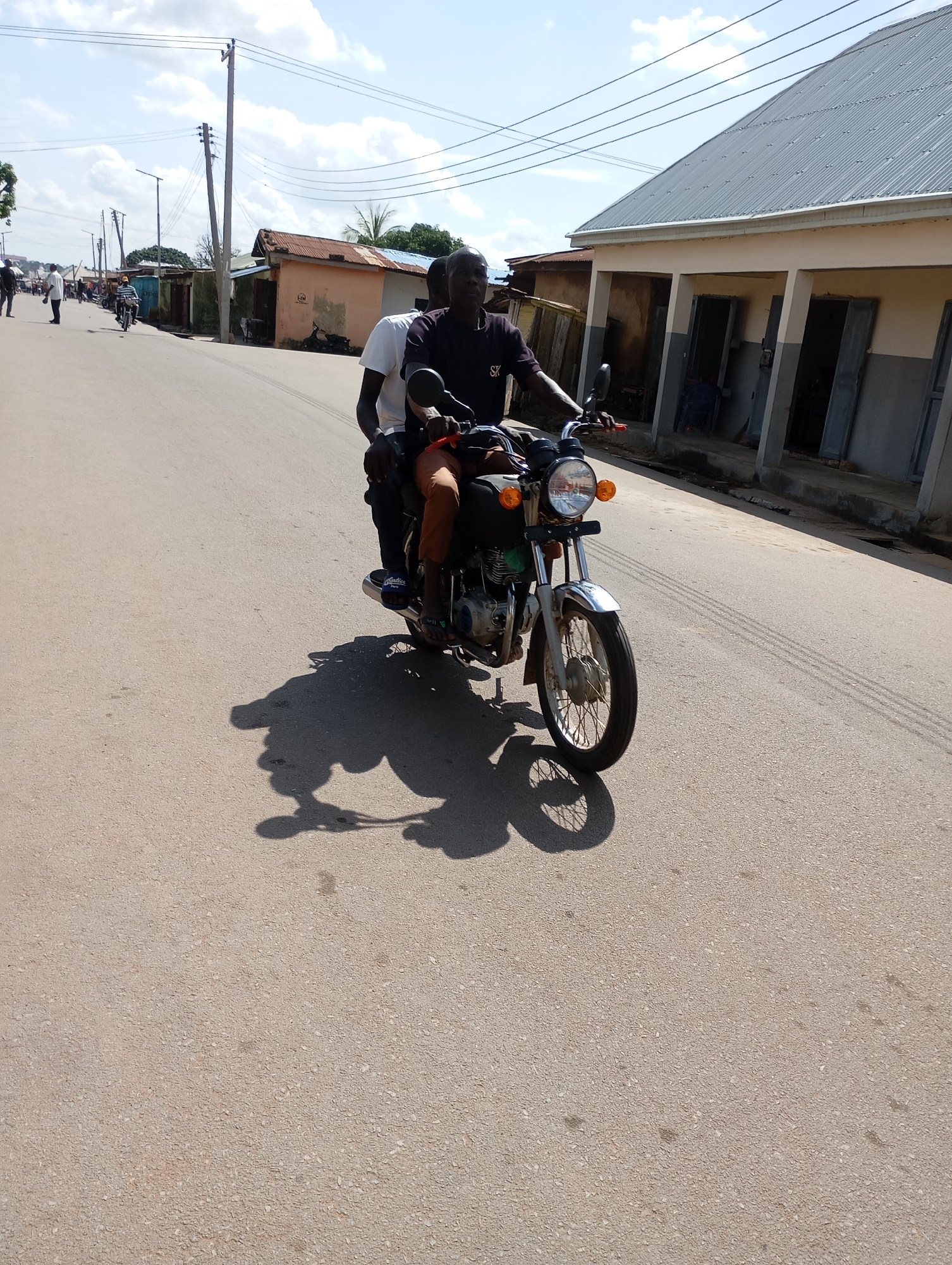 Two Black males are riding a motorcycle on a paved road in Shendam, Nigeria. The rider in front is wearing a black t-shirt with "SX" printed on the left chest and dark pants. The passenger behind him is wearing a white t-shirt and tan pants. The motorcycle is a dark color with a silver front fender and chrome handlebars. The scene is illuminated by bright daylight under a partially cloudy sky. Buildings with corrugated metal roofs and some open doorways are visible along the right side of the road. Utility poles and overhead wires are present. The road surface shows tire marks and shadows cast by the motorcycle and its riders.