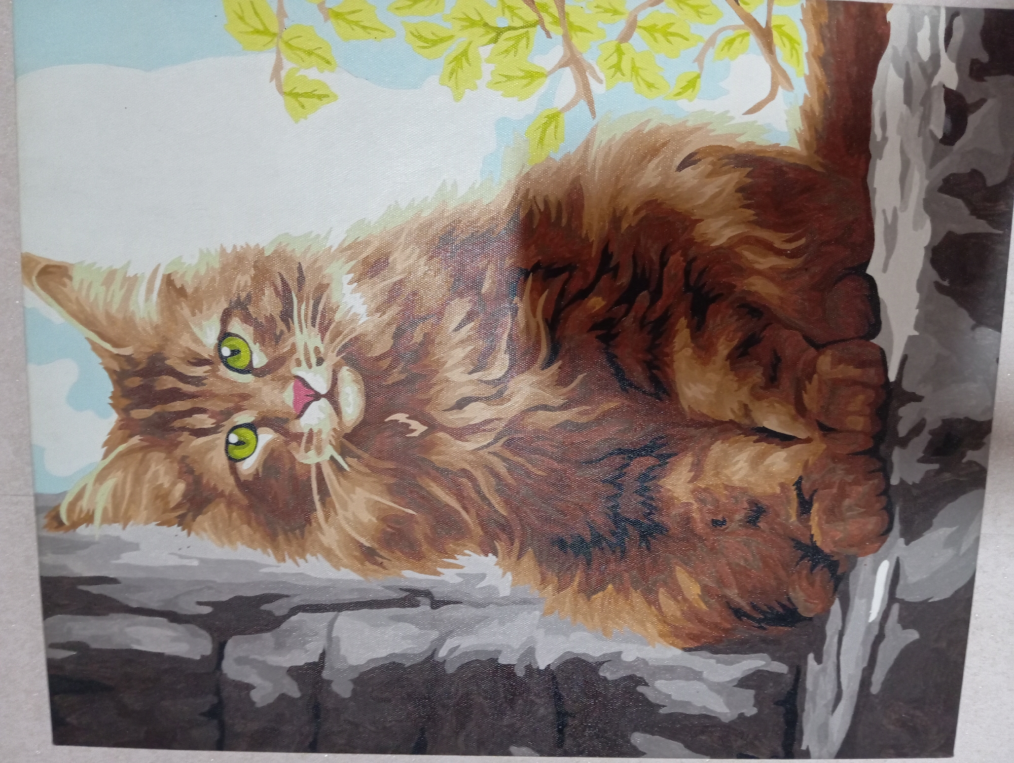 This is a painting of a fluffy, long-haired brown cat with bright green eyes and a pink nose. The cat is sitting on a ledge, with a rough, gray stone wall behind it on the left side of the image. To the right, there is a light blue sky with some stylized yellow-green leaves on branches. The painting appears to be a "paint by numbers" style artwork, given the distinct brush strokes and the somewhat stylized rendering of the cat and its surroundings. There are no people or any indication of a specific location within Barnaul, Russia.