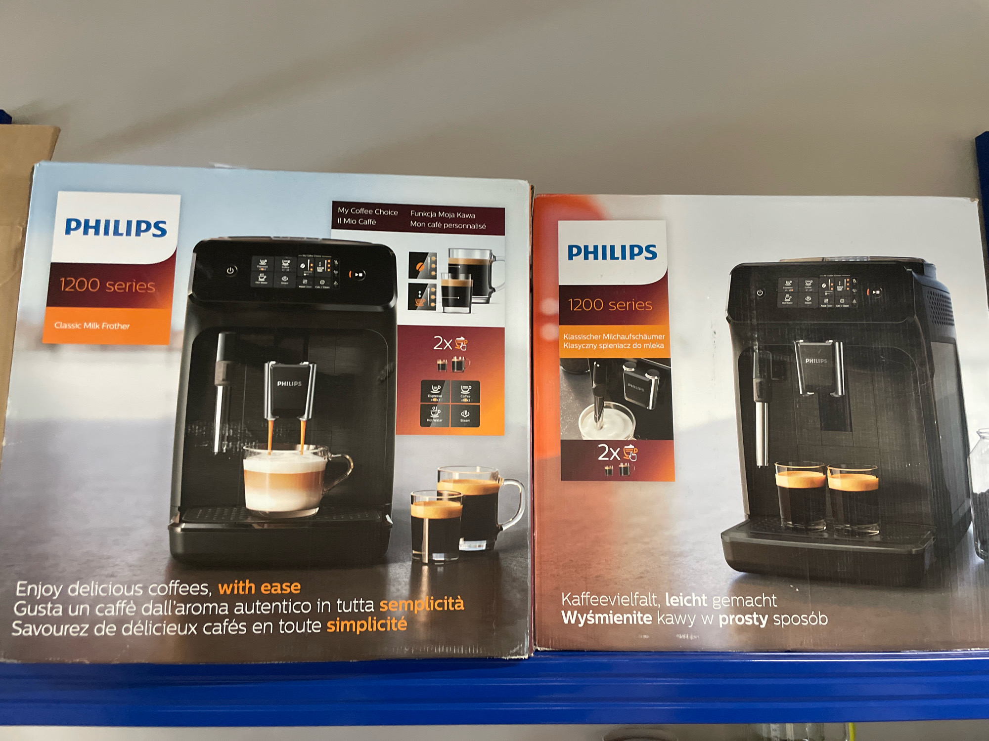 The image shows the packaging for two Philips 1200 series coffee machines, displayed side-by-side on a blue shelf. The left packaging features a black coffee machine with a glass of frothed coffee, and text in English, Italian, and French. The right packaging also displays a black coffee machine, this time with two shots of espresso in glasses, and text in German, Polish, and English. The background suggests an indoor retail environment, likely a store. There are no people visible in the image. The location is not specified, but the overall setting is consistent with a retail display in a store.