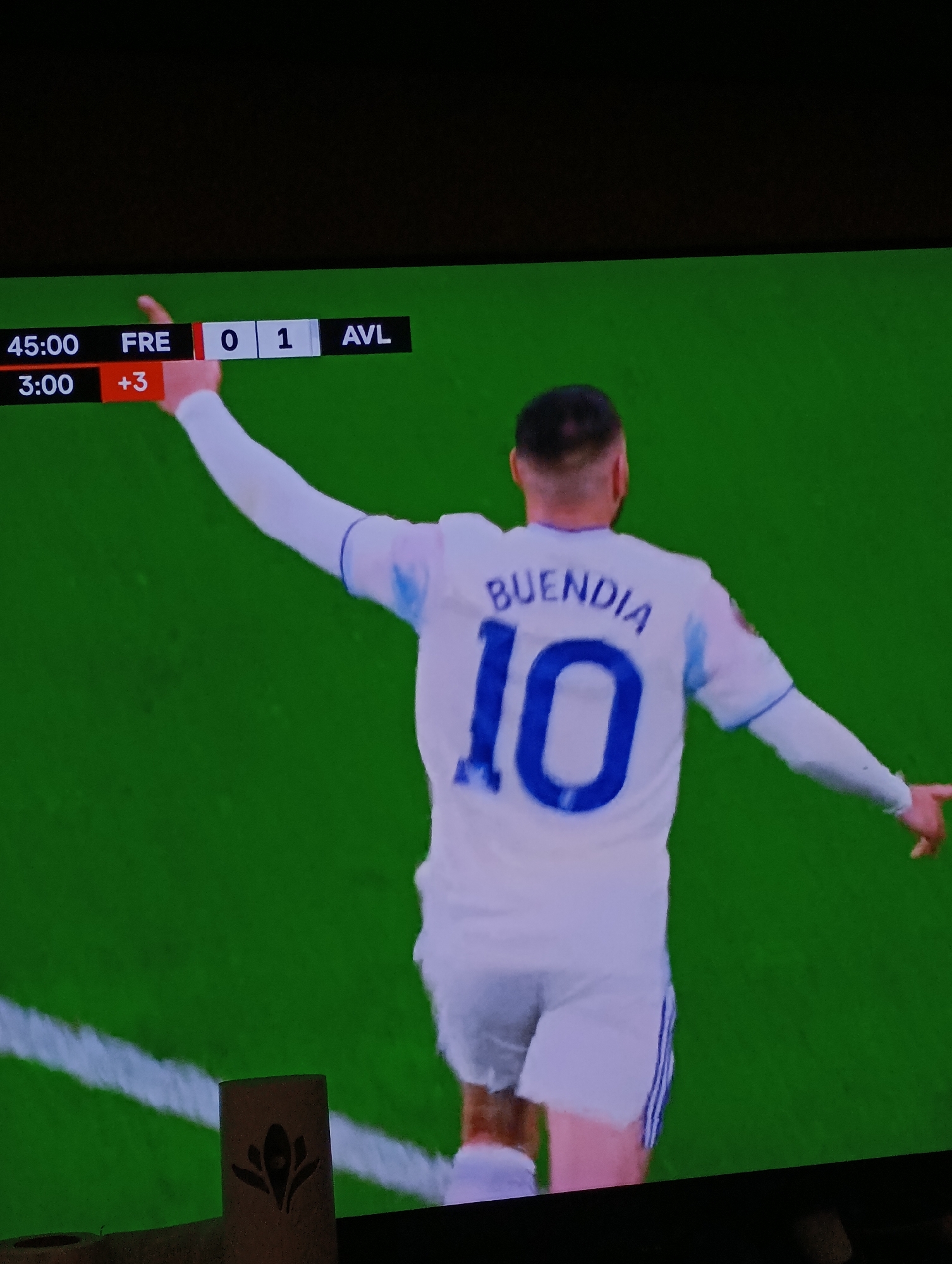 The image shows a soccer (football) match in progress, viewed on a television screen. The score is Freiburg (FRE) 0, Aston Villa (AVL) 1. The game clock reads 45:00 +3. The player on screen is wearing a white jersey with the name "BUENDIA" and the number "10" on the back. He is raising his arms in what appears to be a celebration. The background is a bright green soccer field. The lower left corner of the image also shows a part of what seems like a small wooden structure with a leaf-shaped design. There is no indication of Plovdiv, Bulgaria in this image.