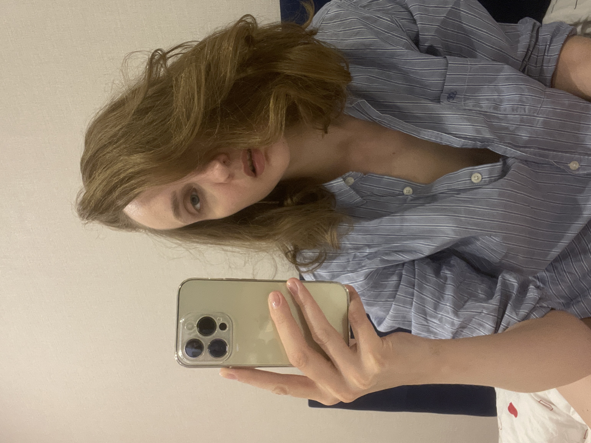 This media file, captured in Podshivalovo, Russia, features a person, likely a young woman, taking a selfie while reclining indoors. The image is oriented with the subject's head to the left and body extending to the right.
The woman has long, light brown or auburn hair that appears wavy and somewhat tousled. She has fair skin, light-colored eyes (appearing blue or gray), and her lips are slightly parted, giving her a neutral to contemplative expression as she looks directly into the camera.
She is dressed in a light blue and white striped button-down shirt, which is partially unbuttoned, revealing a portion of her chest. In her right hand, she holds a light-colored smartphone, resembling an iPhone Pro Max with its distinctive three-lens camera array, encased in a clear cover. Her fingers are visible, and her nails are painted with a light, natural-toned polish.
The setting appears to be an indoor room, possibly a bedroom or living area. A plain, light-colored wall forms the background for most of the image. Behind her head and upper body, a dark blue fabric, likely a pillow or blanket, is visible. In the bottom right corner of the frame, a white sheet or blanket with a subtle red pattern can be seen. The lighting is bright and even, suggesting either daylight indoors or strong artificial illumination. There is no visible text in the image.