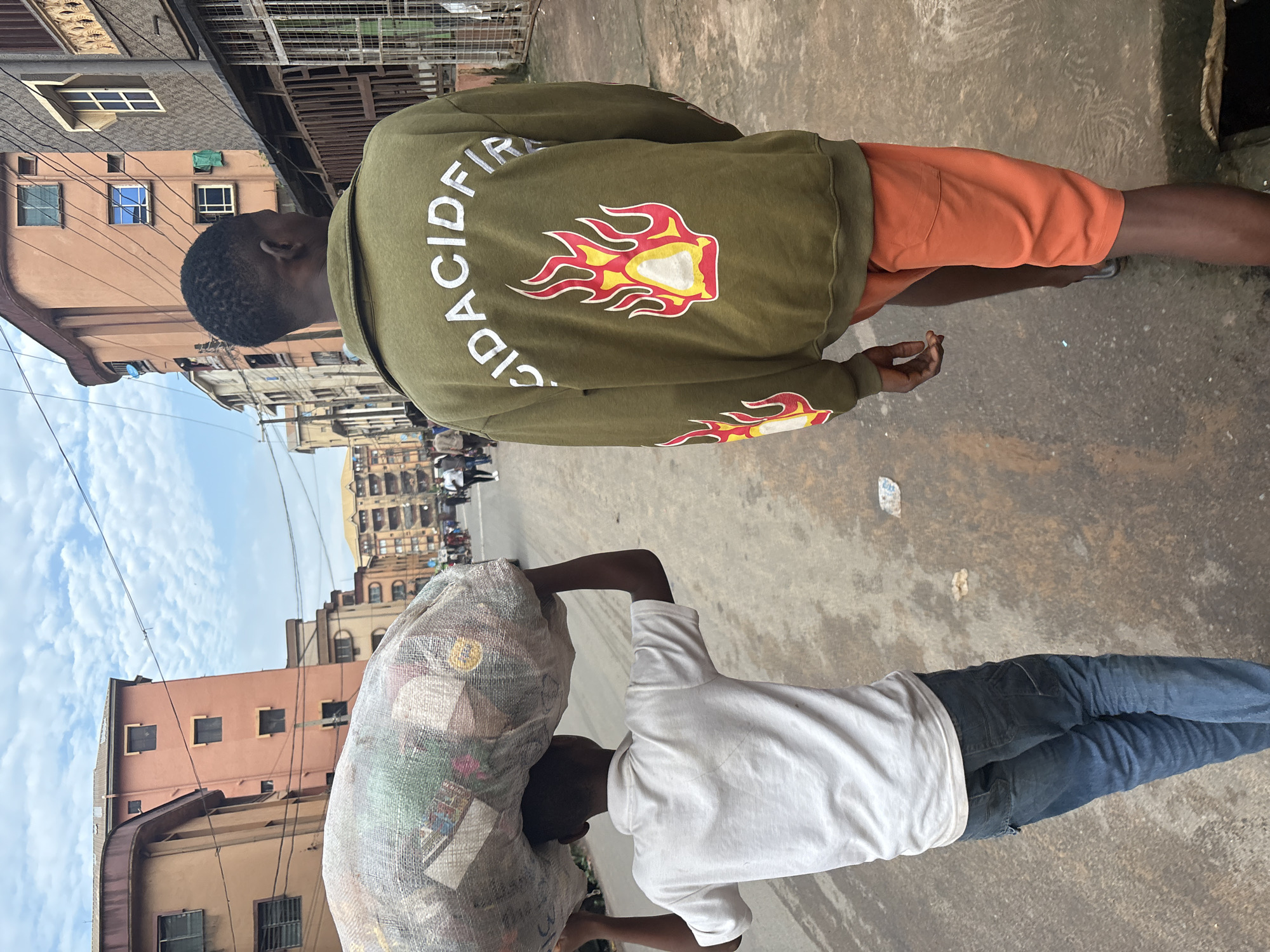 In Onitsha, Nigeria, two young men walk down a paved street lined with multi-story buildings. The man on the left, viewed from behind, carries a large, netted bag filled with various items on his head. He is wearing a white polo shirt and blue jeans. The man on the right, also viewed from behind, walks ahead of him. He is wearing a green sweatshirt with a graphic of flames and the words "ACIDFIRE" in white lettering on the back, and orange shorts. The street is a typical urban environment with some debris visible on the ground. The sky is partly cloudy.