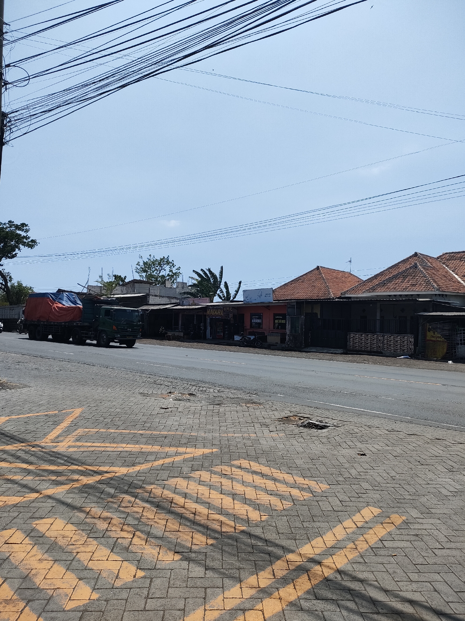 A daytime, eye-level, medium shot captures a street scene in Curahdringu, Indonesia. A dark green truck, its cargo covered by an orange and blue tarp, travels on the right side of a paved road. The road is bordered by a sidewalk made of interlocking gray bricks with yellow painted markings.
On the left side of the road, a row of single-story buildings with red tile roofs line the street. These appear to be shops or small businesses, with signs visible in Indonesian. One sign reads "ANAKA". The architecture suggests a tropical or sub-tropical climate.
The sky is a pale blue with a few faint clouds. Overhead, a dense network of black electrical wires crisscrosses the frame, obscuring parts of the sky and the tops of the buildings. The overall impression is one of a moderately busy commercial street in a developing urban area.