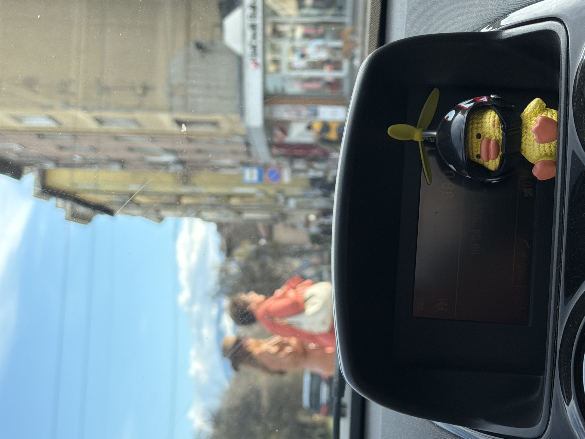 The image depicts an interior view from a vehicle in Sofia, Bulgaria. A dashboard-mounted toy duck with a helmet and propeller is visible to the right. The car window reflects a street scene including buildings, the sky, trees, and two people walking, one wearing a red jacket. A building with a white facade is visible in the reflection.