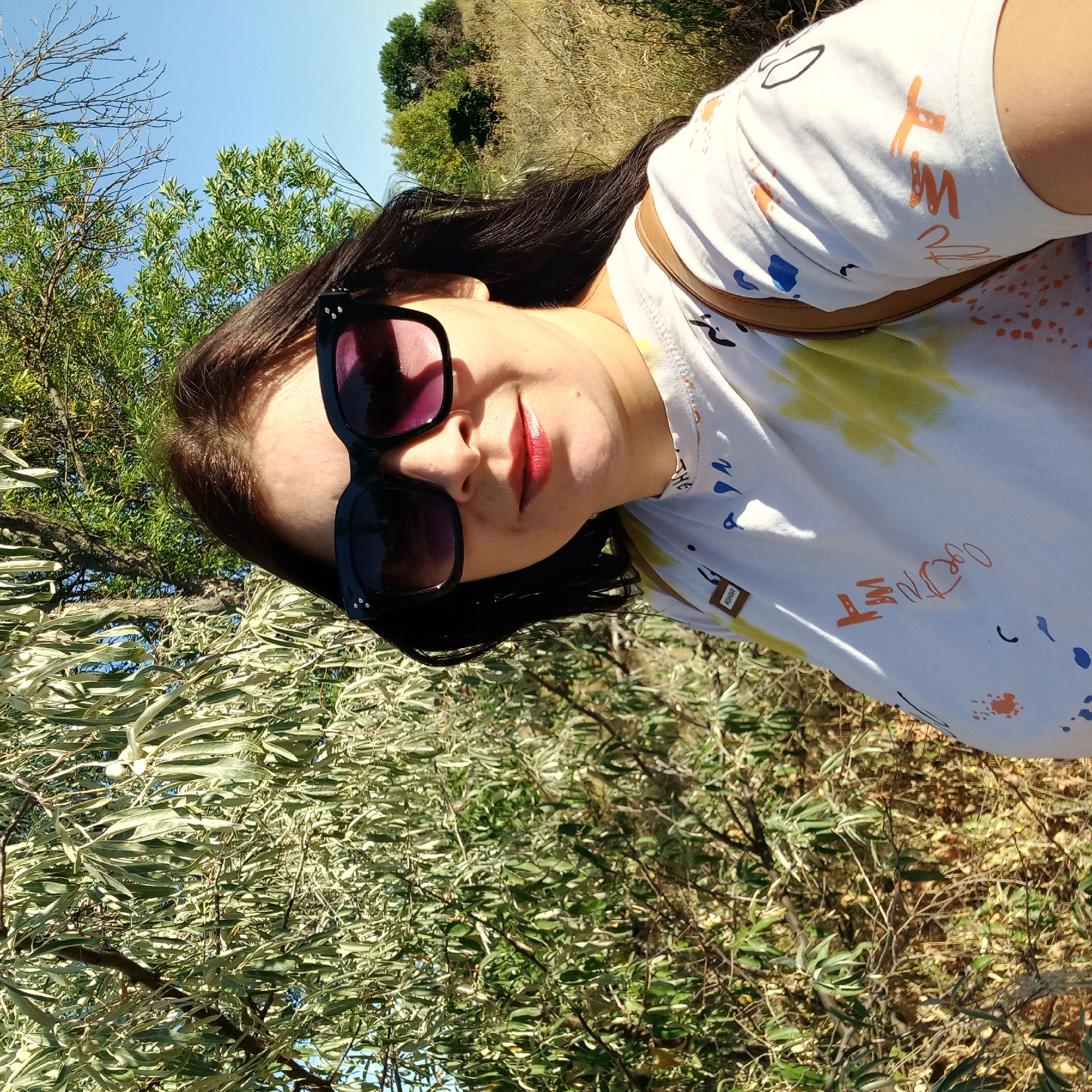 In Berezhynka, Ukraine, a woman with dark hair and sunglasses takes a selfie outdoors. She is wearing a white t-shirt with colorful abstract designs and a brown strap is visible over her shoulder. The background consists of lush green trees and dry grass, with a clear blue sky above. The lighting suggests it is daytime.