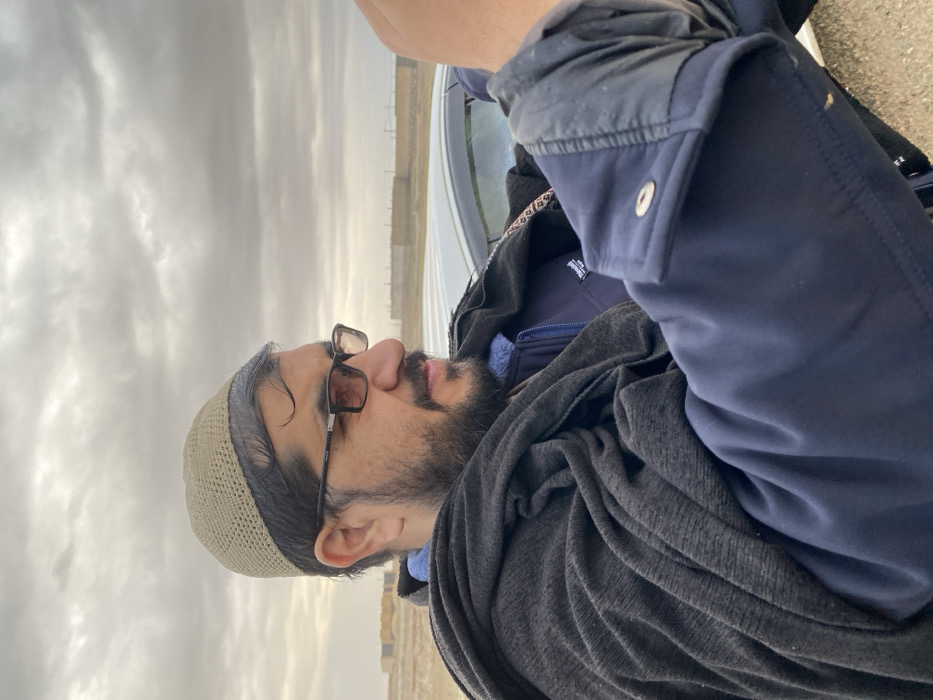 A man with a beard, wearing a tan knit cap and glasses, looks to the left. He's dressed in a navy jacket over a blue shirt, with a gray scarf draped around his shoulders. Part of a white vehicle is visible near the top of the frame. The sky overhead is cloudy, suggesting an overcast day. In the distance, there are structures that appear to be alongside a road.