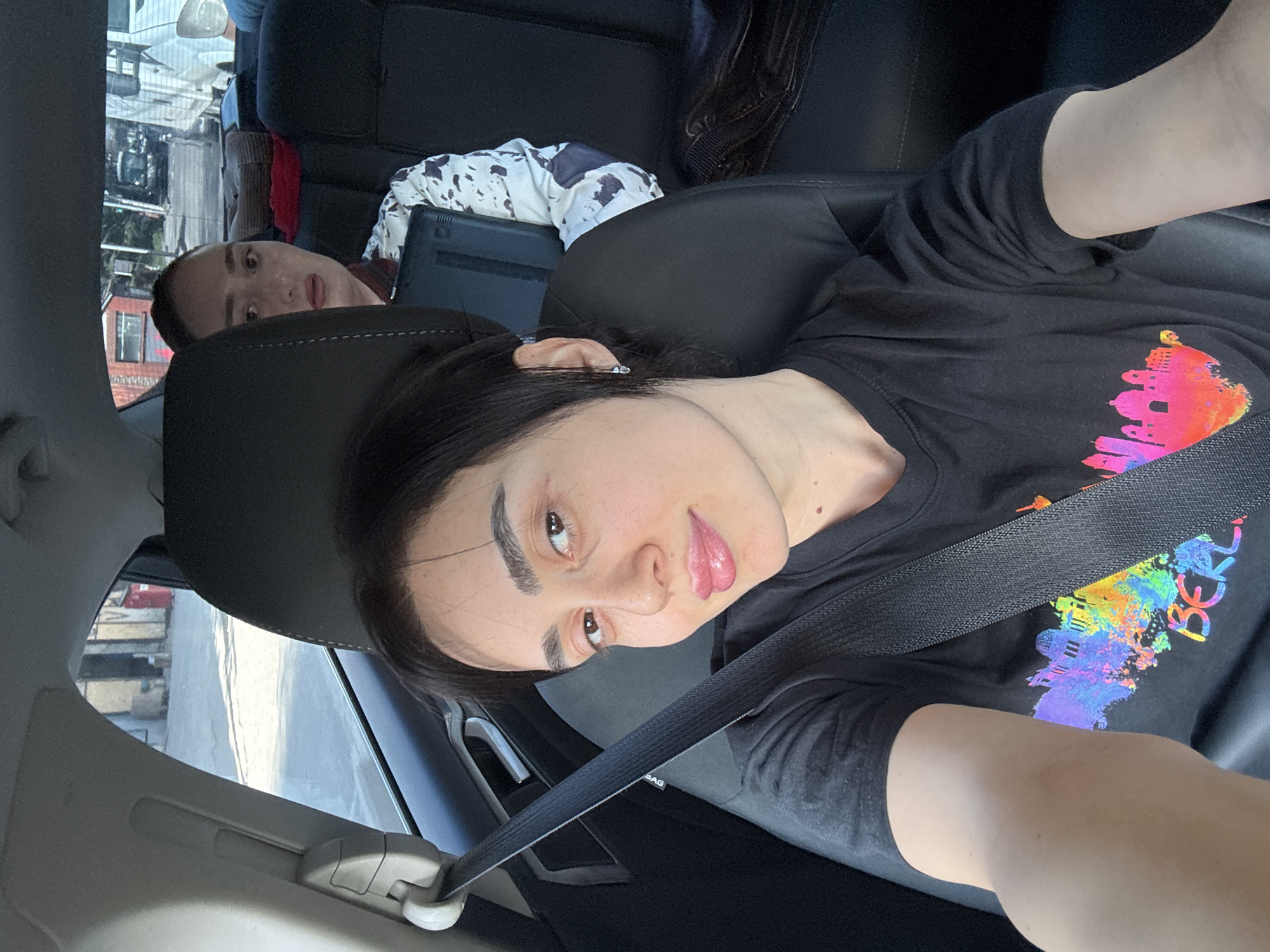 A photograph depicts a woman wearing a black T-shirt with a colorful design on the front and a seatbelt fastened across her chest, sitting in the front seat of a car. Another person is visible in the back seat, partially obscured by the front seat headrest, holding a laptop. The background outside the car window suggests an urban environment, possibly Tbilisi, Georgia. The image has been rotated for an unclear reason.