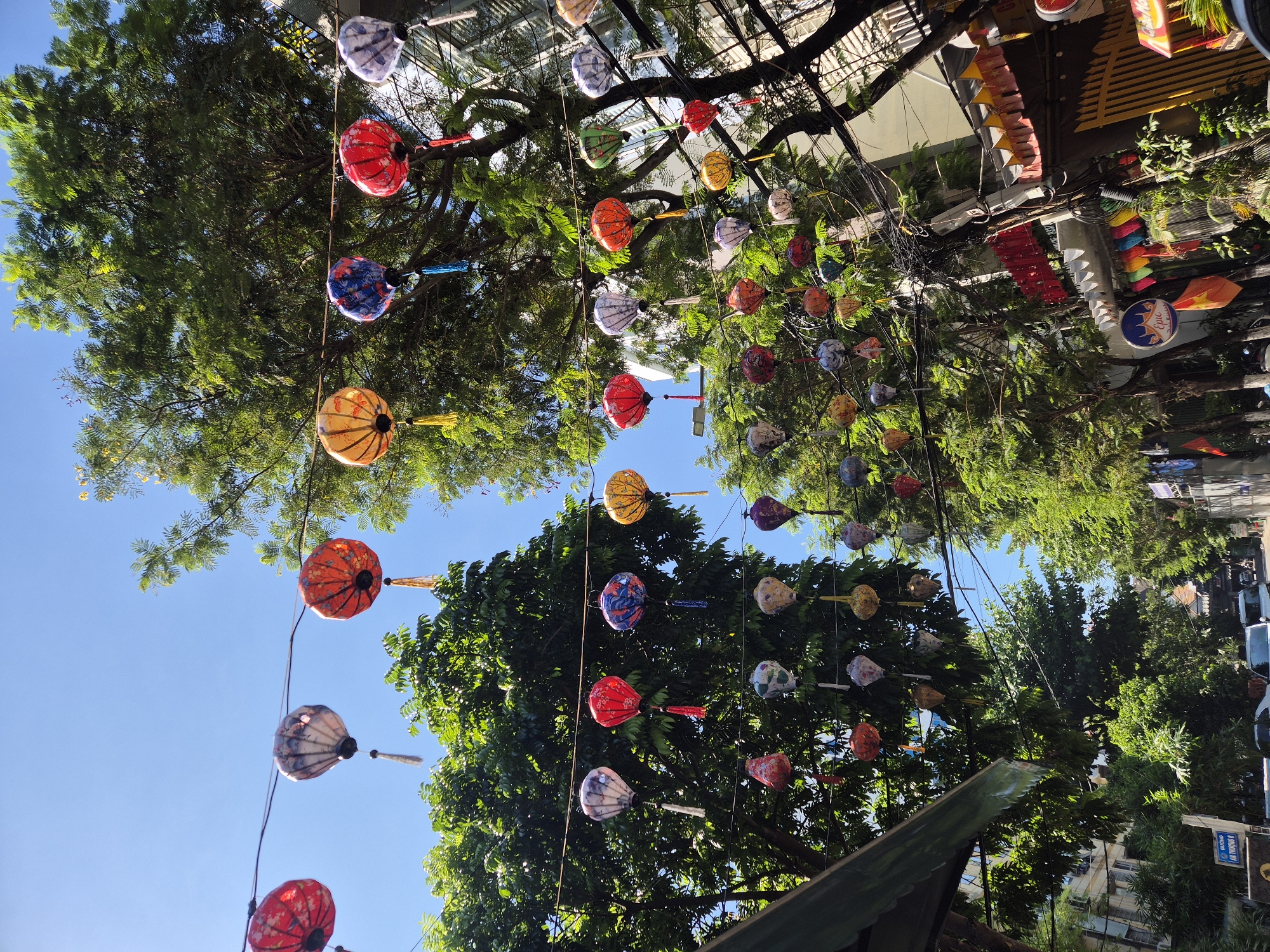 The image depicts a street scene in Da Nang, Vietnam, adorned with numerous colorful lanterns hanging from trees and wires. The lanterns are of various designs and colors, including red, orange, yellow, blue, and white, with some featuring intricate patterns. They are suspended at different heights, creating a festive and vibrant atmosphere.
The scene is set outdoors, with lush green trees and a clear blue sky visible. The sun appears to be high in the sky, indicating it is likely daytime, perhaps late morning or early afternoon, given the bright light and clear shadows. The presence of buildings and what appear to be parked vehicles in the background suggest an urban environment. The overall impression is one of a lively and decorative street, possibly for a festival or as a permanent display to enhance the visual appeal of the area. The style of the lanterns and the surrounding vegetation are indicative of a Southeast Asian location.
There are no people actively visible in the frame, but the scene implies human activity through the decorative elements and the urban setting. The weather appears to be clear and sunny. There is some text visible on a blue circular sign with "Epic" written on it, and another sign with Vietnamese text "TRƯỜNG THPT LÊ TRUNG TÍN". The image conveys a cheerful and celebratory mood due to the abundance of colorful lanterns.