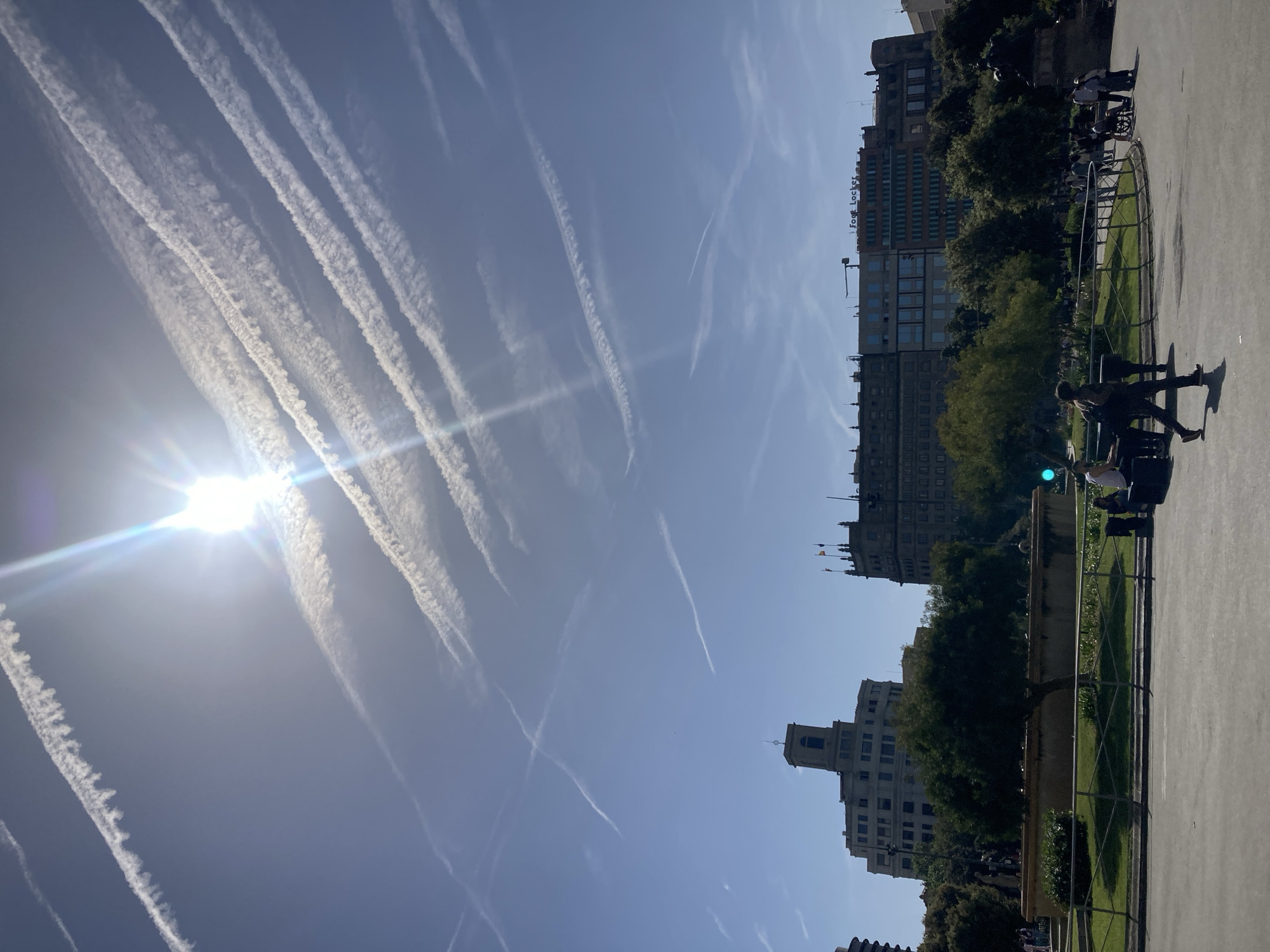 The image depicts a sunny daytime scene in what appears to be an urban park or plaza, with a clear blue sky filled with numerous white contrails from airplanes. The bright sun, accompanied by a starburst lens flare, is prominent in the upper left of the frame.
In the mid-ground and background, a series of multi-story buildings line the perimeter of the public space. One large building distinctly features the "Foot Locker" logo prominently on its facade. Other buildings show various architectural styles, contributing to an urban cityscape. Below the buildings, a strip of green trees and manicured foliage separates them from the foreground.
The foreground consists of a wide, light-colored paved area, possibly a walkway or plaza, with metal barriers and low walls defining grassy sections. Several individuals are visible in the mid-ground and foreground. Some people are walking, casting shadows on the pavement, while others appear to be standing or resting. One person is seen pushing a stroller or wheelchair. A lamppost with a green light is also present in the scene. The overall impression is a vibrant, clear day in a city environment.