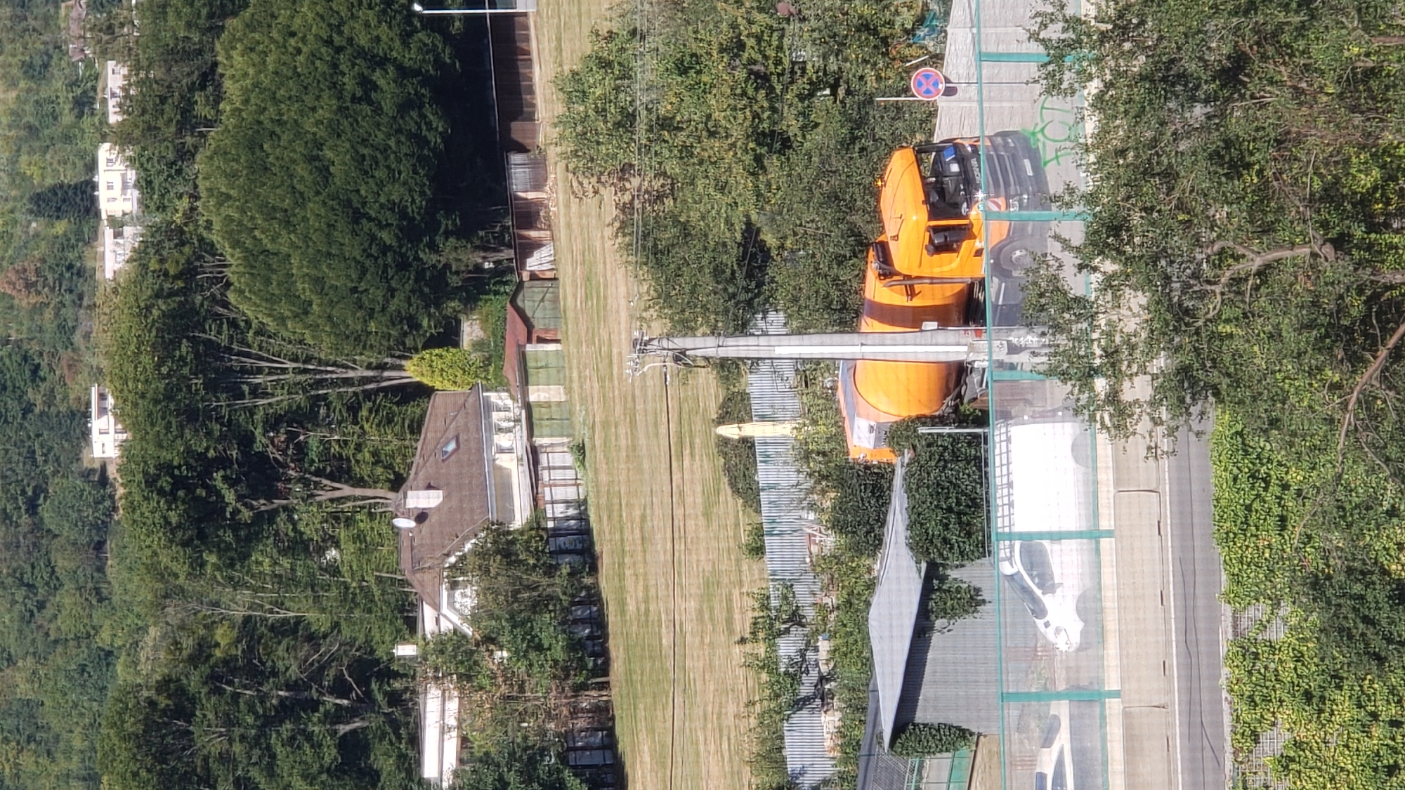 The image shows an elevated view of a residential and industrial area. In the foreground, an orange truck with a cylindrical tank is parked. A white van with a ladder rack on top is visible to the left of the truck, behind a green fence. A concrete barrier runs along the road beneath the vehicles. In the middle ground, there are several sheds and a corrugated metal structure with ivy growing on it. A large, dry, grassy field is visible behind these structures, leading up to a cluster of trees. In the background, several white buildings with red roofs are nestled among dense green trees on a hillside. The sky is not visible in the frame. The overall impression is of a semi-urban landscape with a mix of residential and utilitarian structures.