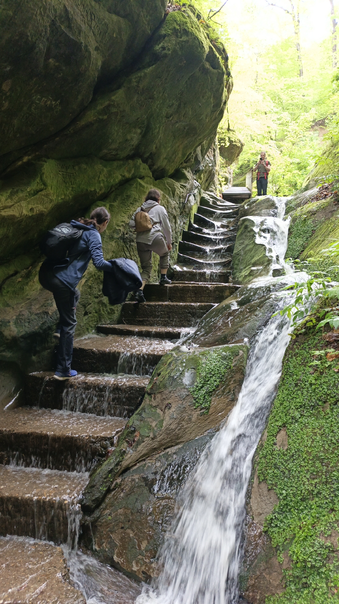 Two women are climbing a stone staircase that is part of a waterfall in a lush, green environment. A third person stands on a bridge at the top of the stairs. The staircase is built into a rock face covered in moss and plant life. Water is flowing down the stairs, creating a cascade effect. The location is in Goryachiy Klyuch, Russia.