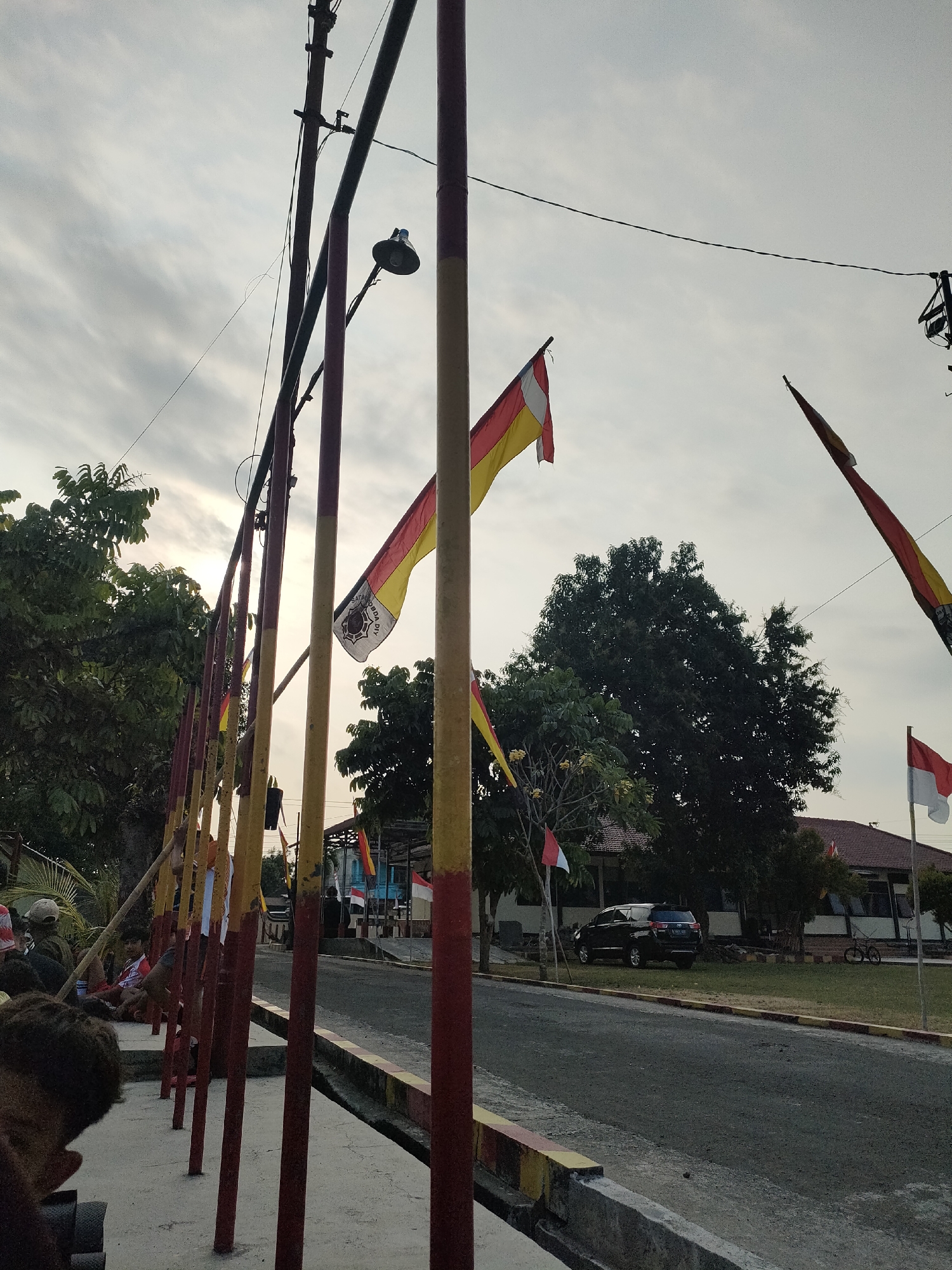The image captures an outdoor scene with a road and a row of red and yellow striped poles, likely part of a temporary barrier or decoration, in the foreground. Several flags are visible, including a red and white Indonesian flag and other flags with yellow and red bands. Trees and a building with a tiled roof are present in the background. A black car is parked on the grass near the building, and a bicycle is visible in the distance. The sky is overcast with some light filtering through. The setting appears to be a public area, possibly a street or a park, in Banguntapan, Indonesia, given the presence of Indonesian flags. Several people are seated or standing behind the poles on the left side of the image, though their faces are not clearly visible. One flag, prominently displayed on the right side, has a crest or emblem on it.