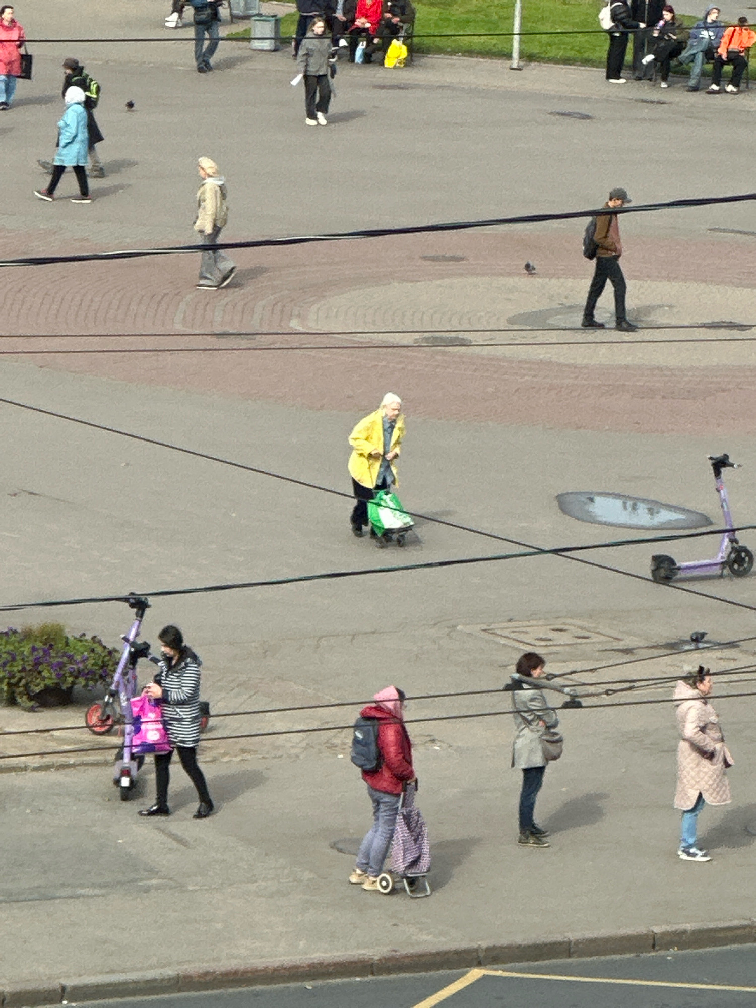 The image depicts a wide-angle street-level view, likely in Saint Petersburg, Russia, with several pedestrians and electric scooters present on an asphalt-paved area adjacent to a sidewalk. The scene is divided by dark horizontal power or cable lines, creating distinct layers of activity.
In the upper portion of the image, several individuals are walking on the paved area. A person in a light blue jacket walks towards the left, while another in a dark jacket is behind them. Further to the right, a person in a grey jacket walks towards the left, carrying a white bag. In the distance, other figures are visible, some with shopping bags.
The middle section of the image features an elderly woman in a bright yellow jacket pushing a green shopping cart with wheels. She is walking towards the right. To her right, a purple electric scooter is parked.
In the lower section, a woman in a striped coat stands next to a purple electric scooter, holding a pink shopping bag. Further to the right, a woman in a red jacket with a backpack is walking towards the right, pulling a wheeled cart. Next to her, a woman in a grey coat stands, and to her right, a woman in a patterned beige coat stands. A portion of a building facade with a window is visible in the far background. The overall environment suggests an urban public space.