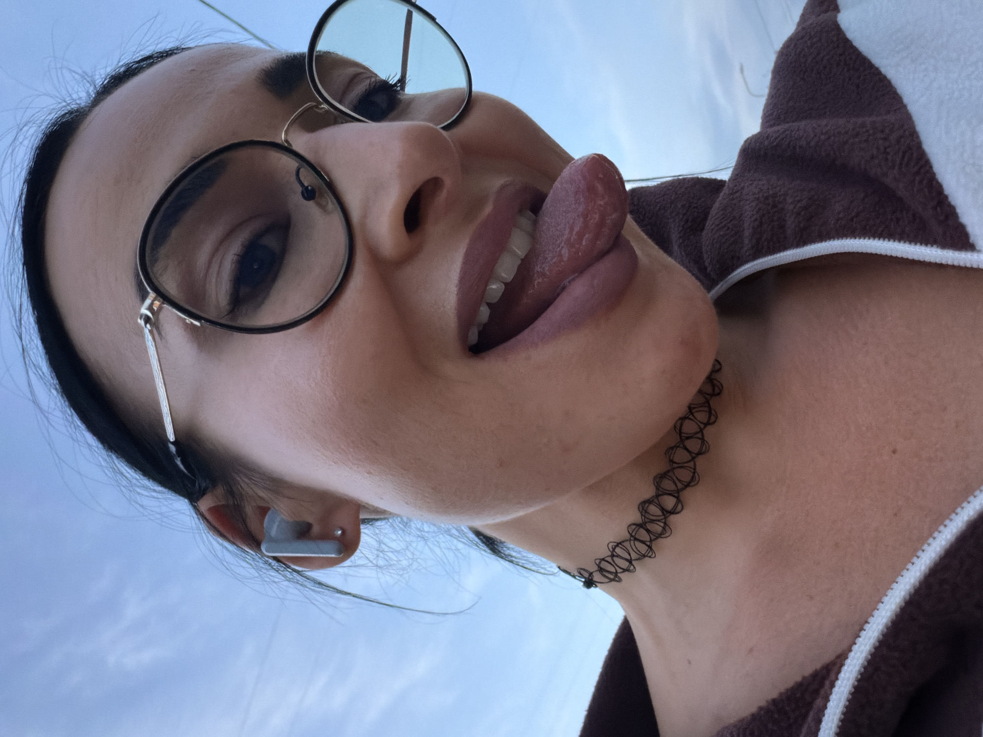 A close-up, low-angle selfie captures a person, likely female, looking upwards towards the camera against a clear, light blue sky, indicative of daytime. The subject wears round, metal-framed glasses that are slightly pushed up. A grey wireless earbud is visible in their left ear.
The person has a playful expression, with their tongue partially extended. They are wearing dark lipstick, and their teeth are visible. Around their neck, a black, elastic, spiral-patterned choker necklace is worn. The visible clothing includes a brown and white hooded jacket, possibly fleece, with a white zipper detail on the right side of the frame. Dark hair is pulled back, with some loose strands around the ears and forehead. The overall mood is lighthearted and confident. There is no visible text in the image. The location context for this image is Saint Petersburg, Russia.