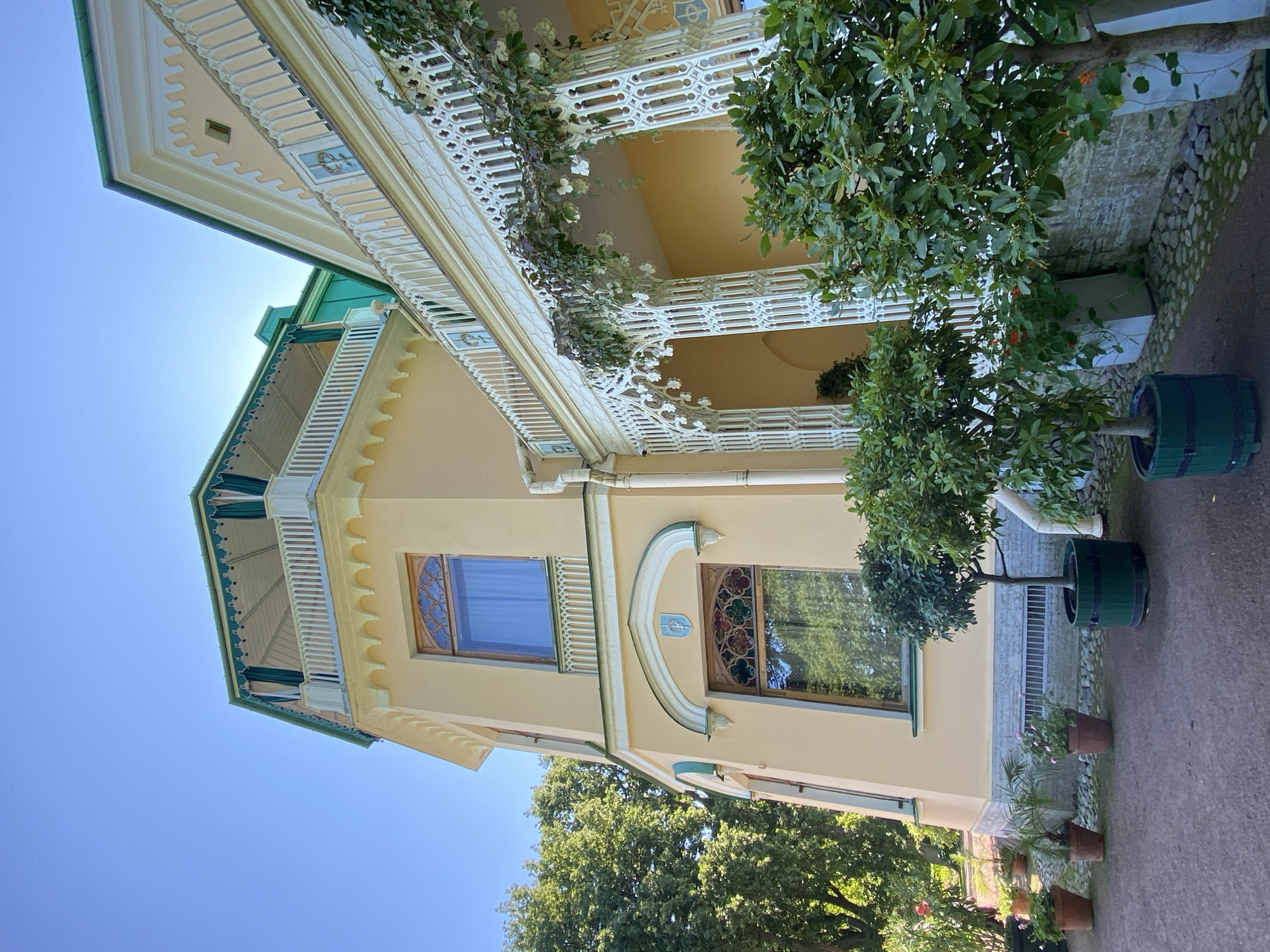 The image captures the facade of a yellow building with green trim, likely part of the Peterhof Palace complex in Peterhof, Russia. The architecture features ornate white decorative elements along the balconies and porch, with intricate latticework supporting the roofline of the porch. Several potted trees and plants are arranged in front of the building, with one larger tree in a dark green planter to the right. The sky is clear and blue, indicating a sunny day. The building has multiple windows, some with decorative stained glass. The ground in front of the building is paved with cobblestones. There are no people visible in the image.