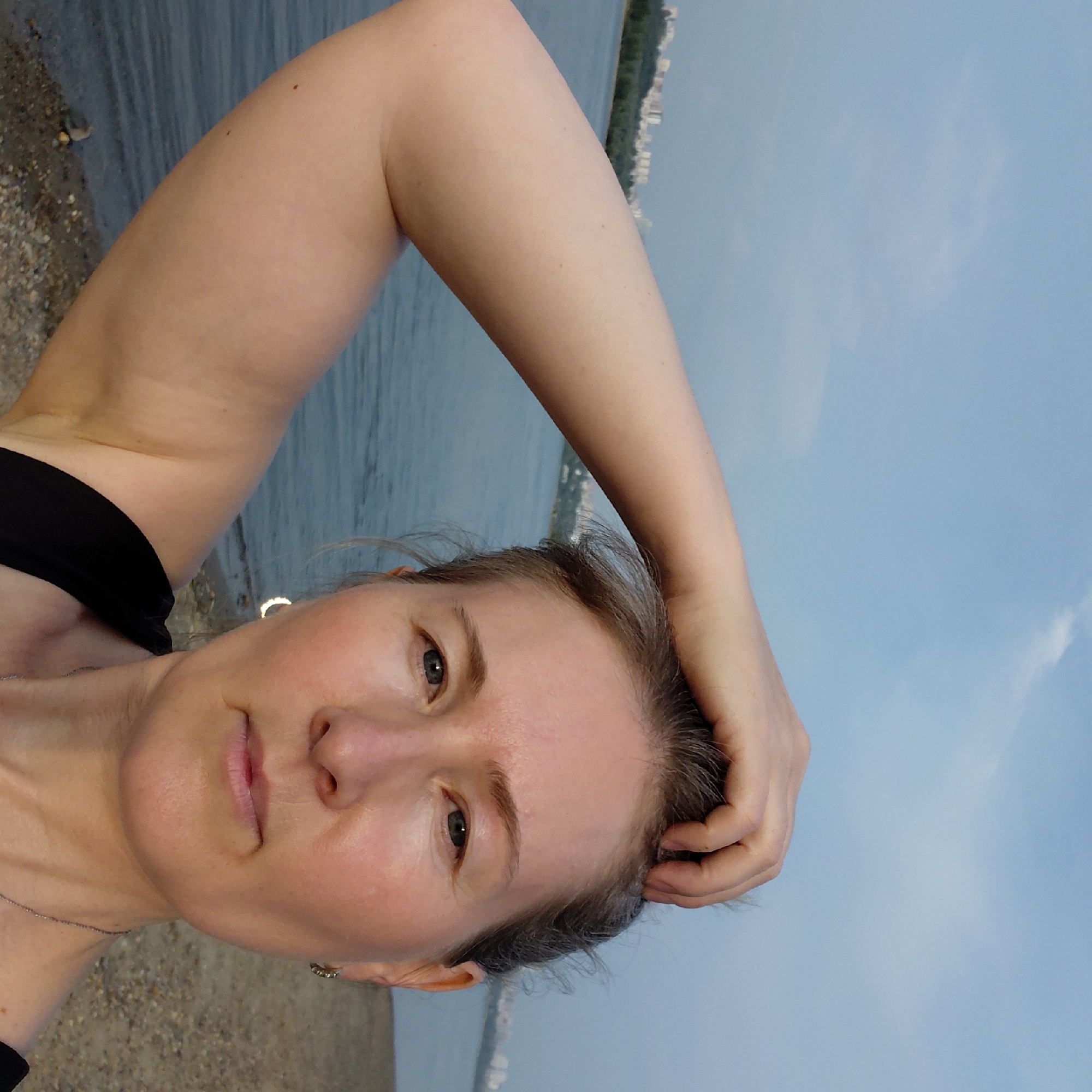 The image is a selfie taken outdoors with a body of water and a city skyline in the background. The subject is a woman with her arm raised, touching her head. She is wearing a black top and a necklace. The environment appears to be a beach or waterfront area with a sandy shore. The city skyline in the background suggests an urban setting. Based on the presence of buildings that resemble those found in Novosibirsk, it is possible this photo was taken in that city, perhaps along the Ob River, which runs through Novosibirsk. The sky is partly cloudy.