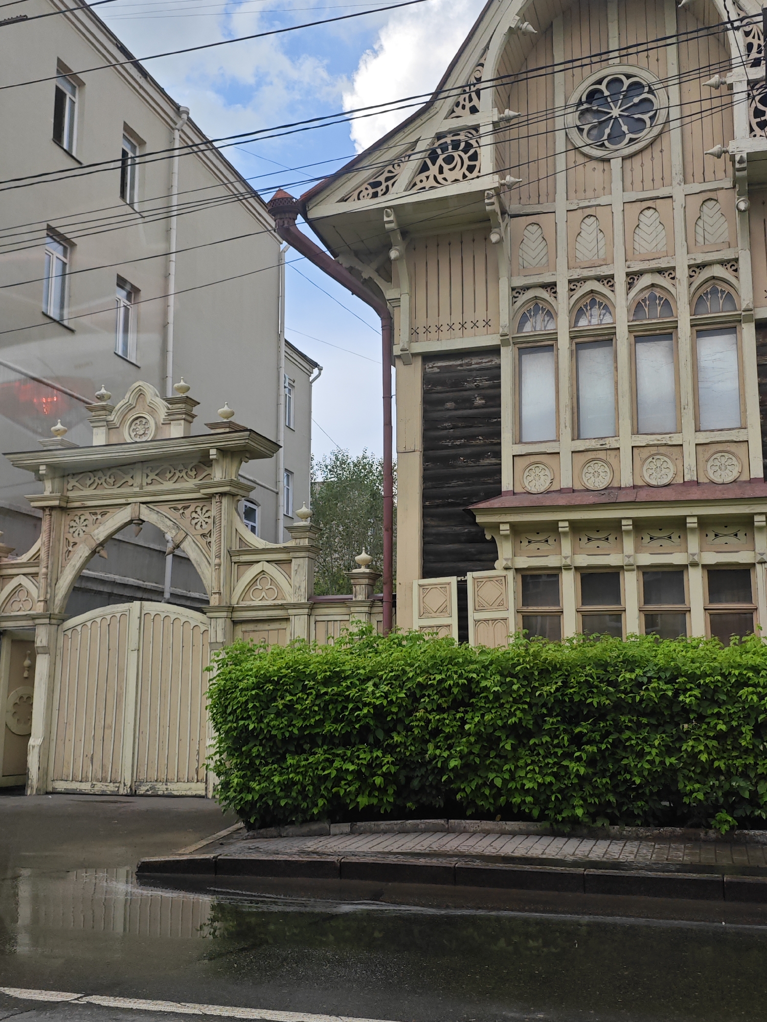 The image captures a view of a street in Krasnoyarsk, Russia, featuring a juxtaposition of two architectural styles. On the right, an ornate, wooden, two-story building with intricate carvings and a round decorative window dominates the frame. This building appears to be older, with a traditional style often seen in historic Russian architecture. To its left, a more modern, grey, multi-story building stands, presenting a simpler, functional design with regularly spaced windows. A tall, ornate wooden gate with detailed latticework and decorative finials is visible in the foreground, leading to what seems to be a courtyard. A lush green hedge lines the sidewalk in front of the gate, and the street in the foreground is wet, reflecting the overcast sky and the surrounding structures, suggesting recent rain. Power lines crisscross the sky, a common urban element. The scene appears to be a quiet street, possibly in a residential or historically significant area of the city.