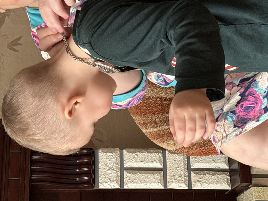 An indoor scene in Pavlovsky, Russia, depicts a young child and a partially visible adult. The child, with blonde hair and wearing a dark green long-sleeved shirt, is shown from the head to the waist, looking downwards. An adult's hands are visible behind the child's neck, fastening a silver-colored chain necklace with a decorative pendant around the child's neck. The adult is wearing a light blue and pink patterned top. On the left, a dark brown wooden structure with fluted carvings stands above a section of wall or furniture faced with light-colored, rectangular, textured blocks resembling stone. A wall with light-colored, textured wallpaper or paint is visible further back. A textured fabric with orange, brown, and beige hues is present in the middle-ground. The lower portion of the image displays a leg, likely belonging to the child, clad in a garment with a white background and a pattern of pink and purple roses.