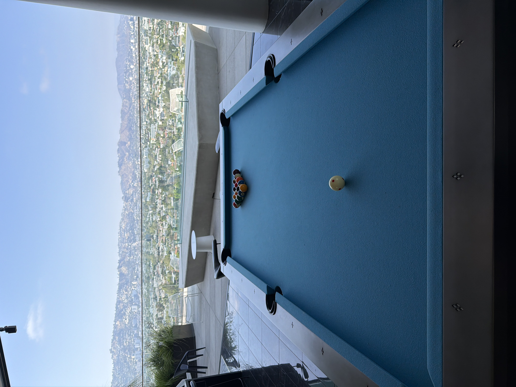 A high-angle, vertically-oriented shot captures a pool table set up on an outdoor patio. The patio offers a scenic view of the city of Los Angeles, California, USA.
The pool table, with a blue felt surface, is fully racked at one end and has a cue ball placed at the opposite end, ready for a game. The table is positioned on a tiled floor, and beyond the patio railing is a vista of the city sprawling across the landscape, with mountains in the background under a clear sky. A small, round white table and patio furniture can be seen to the left of the pool table. The image suggests a luxurious outdoor space designed for entertainment and relaxation, with a stunning view of Los Angeles.