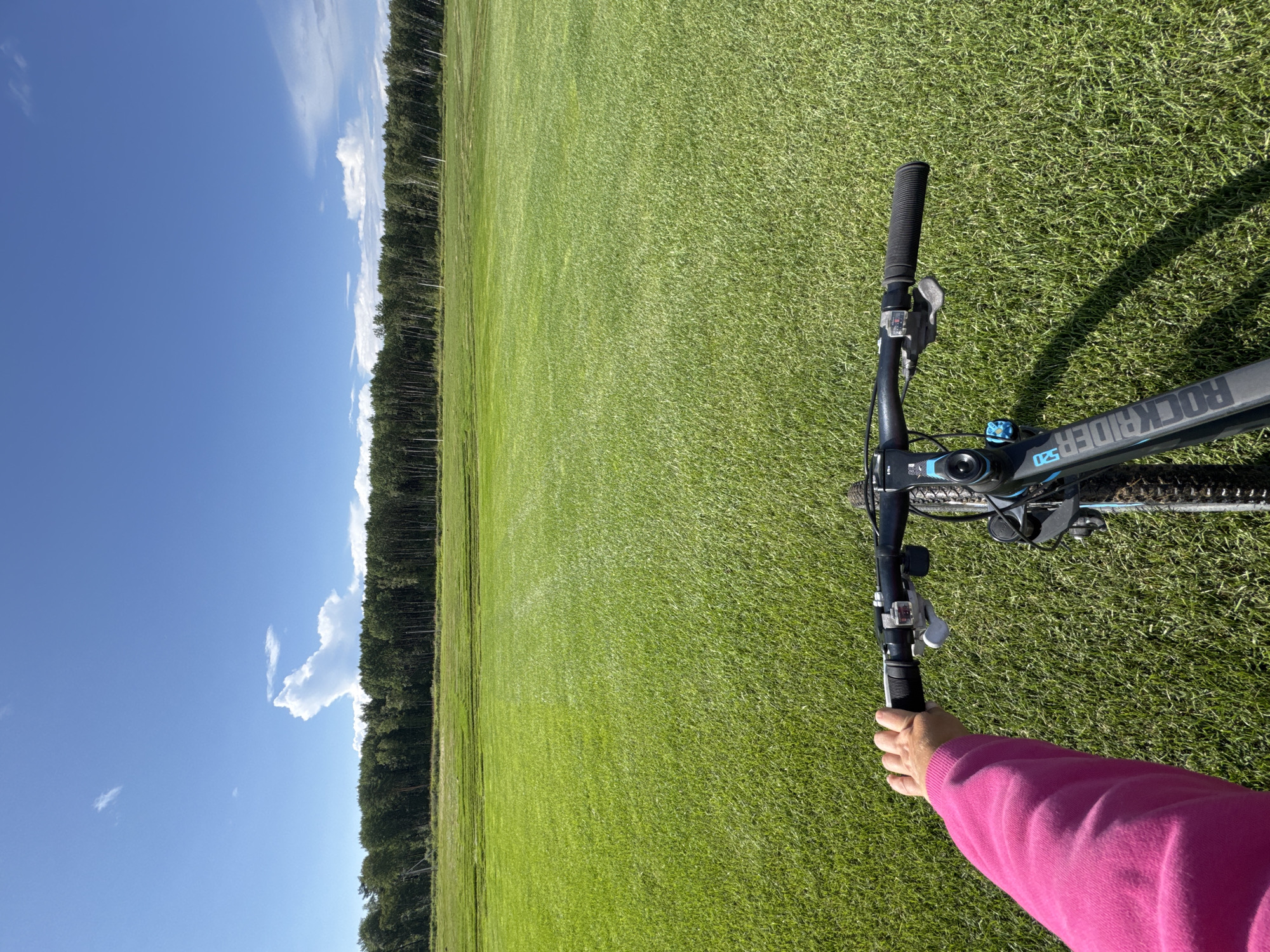A first-person perspective shot captures a person on a bicycle, holding the handlebars with their left hand, wearing a bright pink sweatshirt. The bicycle is black and blue with "ROCKRIDER" branding visible on the frame. The setting is a vast, freshly cut green field under a clear blue sky with scattered white clouds. A dense line of trees borders the far edge of the field. The image appears to be taken in or near Innolovo, Russia, given the context provided.