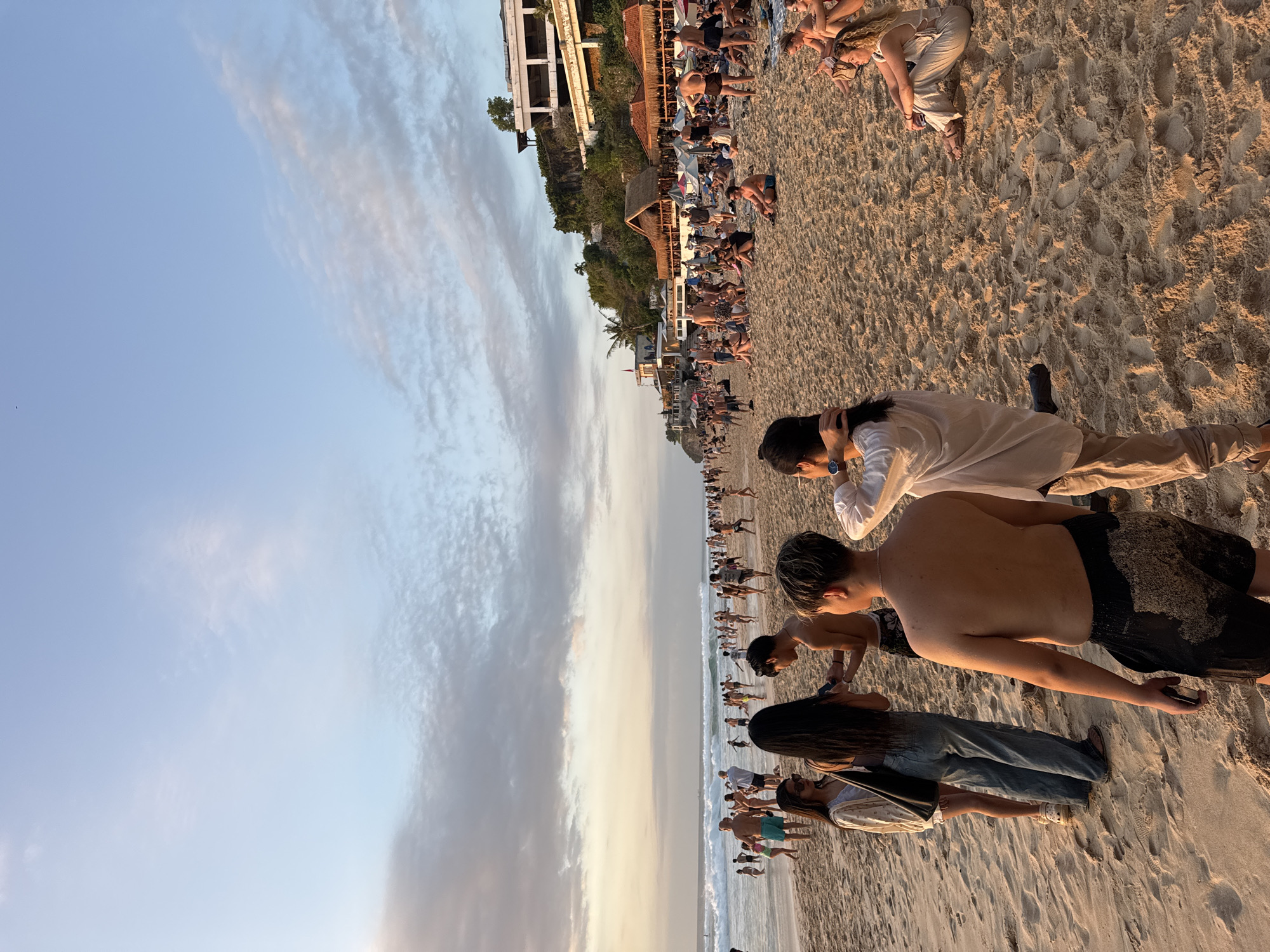 The image captures a crowded beach scene in Kuta Selatan, Indonesia, during what appears to be late afternoon or early evening. The sky is a soft blue with scattered, wispy clouds, tinged with the warm hues of an approaching sunset. The sandy beach is populated with a diverse group of people, many dressed in beachwear.
In the foreground, several individuals are visible. A shirtless young man with dark hair faces away from the camera, wearing black shorts covered in sand. To his left, a young woman with long dark hair stands looking towards him, wearing sunglasses and a light-colored cover-up over a dark top. Next to her, another young woman in blue jeans and a white top is also looking at the man. Further to the right, a group of people are seated on the sand. One woman with blonde hair is wearing a beige outfit and is looking to her left. Another woman to her right is wearing a white bikini top and shorts.
The middle ground of the image shows more beachgoers. Some are walking along the shore, while others are wading in the ocean, which has moderate waves breaking. The ocean appears to extend to the horizon, meeting the sky.
The background reveals a developed area along the coastline, with several buildings and thatched-roof structures that appear to be restaurants or resorts. Umbrellas are scattered across the beach, indicating areas of shade. Palm trees are visible amongst the buildings, consistent with a tropical environment. The overall impression is of a popular tourist destination buzzing with activity and social interaction as people enjoy the beach and the evening light.