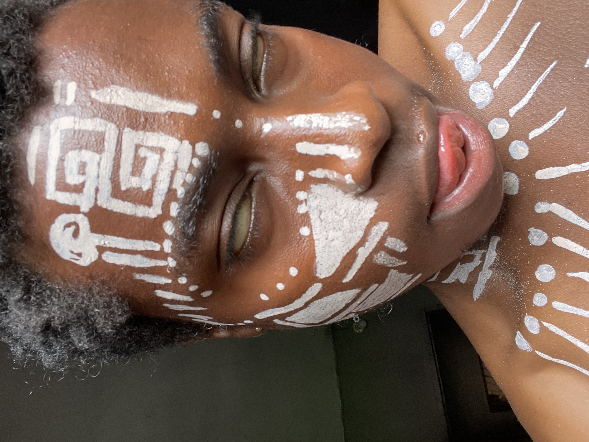A close-up, tilted shot shows a person with dark skin adorned with white face and body paint. The paint is applied in geometric patterns on the forehead and cheeks, and radial patterns of lines and dots extend onto the chest and shoulder. The person's eyes are closed and their mouth is slightly open, revealing their tongue. The person's hair is short and dark. The background is dark and partially out of focus. There are no obvious clues to indicate the location is Onitsha, Nigeria.