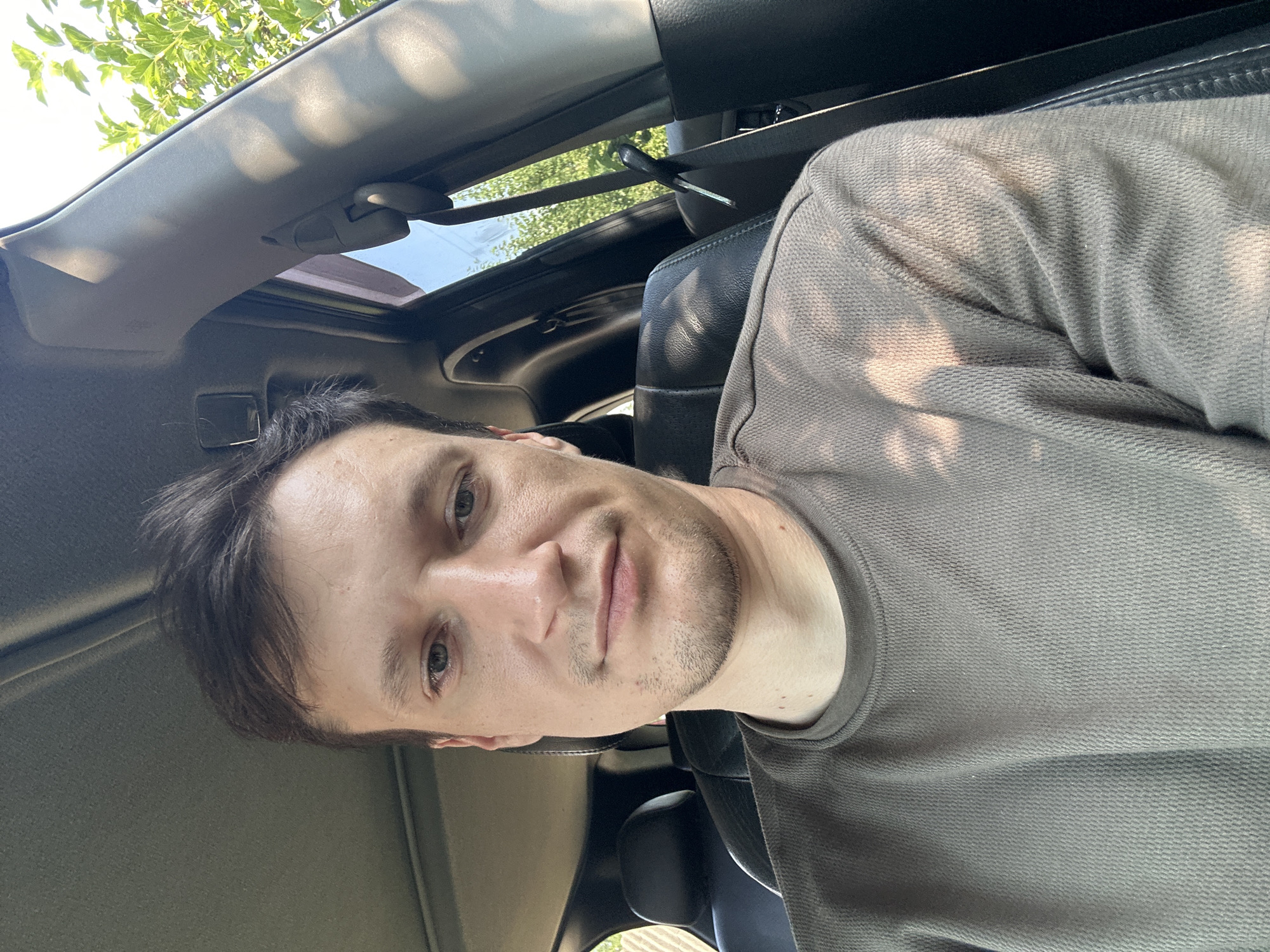 A medium shot captures a selfie of a young adult male inside a vehicle, taken from the chest up. The man has short, dark brown hair, light skin, and a subtle stubble. He is wearing a plain, dark brown long-sleeved shirt. His expression is neutral, and he is looking directly at the camera. The interior of the car is visible, with black leather seats and a sunroof. Sunlight filters through the trees outside, casting dappled shadows on the man and the car's interior. The background includes greenery from trees, suggesting an outdoor setting. The location is identified as Shatava, Ukraine.