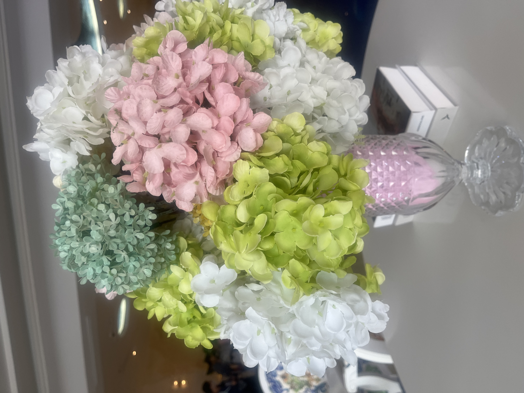 An overhead shot displays a bouquet of artificial hydrangeas in pastel shades of green, pink, and white. The flowers are arranged in a clear, ornate glass vase with a diamond-cut pattern that glows pink from within. The vase rests on a smooth, light-gray surface, presumably a table. Two white-covered books are stacked to the right of the vase. In the background, out of focus, a dimly lit interior is visible, with some lights reflecting on a ceiling. Two indistinct figures are present in the far background. The scene is likely indoors.