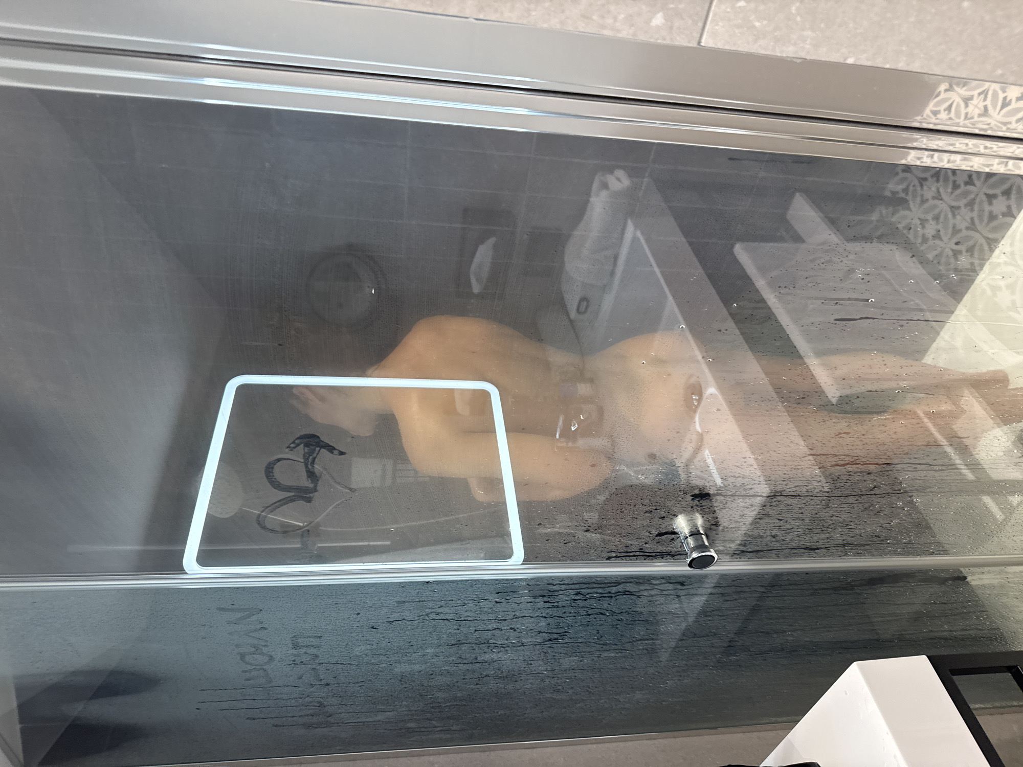 A person is visible inside a steamy glass shower enclosure. The individual appears to be relaxing, possibly lying down or leaning against the shower wall, with their legs and torso partially visible through the fogged glass. A white-bordered, transparent, square-shaped stand or holder, resembling a waterproof tablet or phone mount, is affixed to the shower glass. Dark, squiggly lines or symbols are visible through or on this stand.
The shower glass is covered in condensation and numerous water droplets, indicating the use of warm water. A metallic shower handle or knob is visible on the glass door. Outside the enclosure, light-colored bathroom tiles are visible, with some showcasing a decorative patterned design. Faint, inverted text that appears to read "NYOAN" is scrawled on the lower section of the glass door. The overall scene suggests a private and relaxing moment in a bathroom.