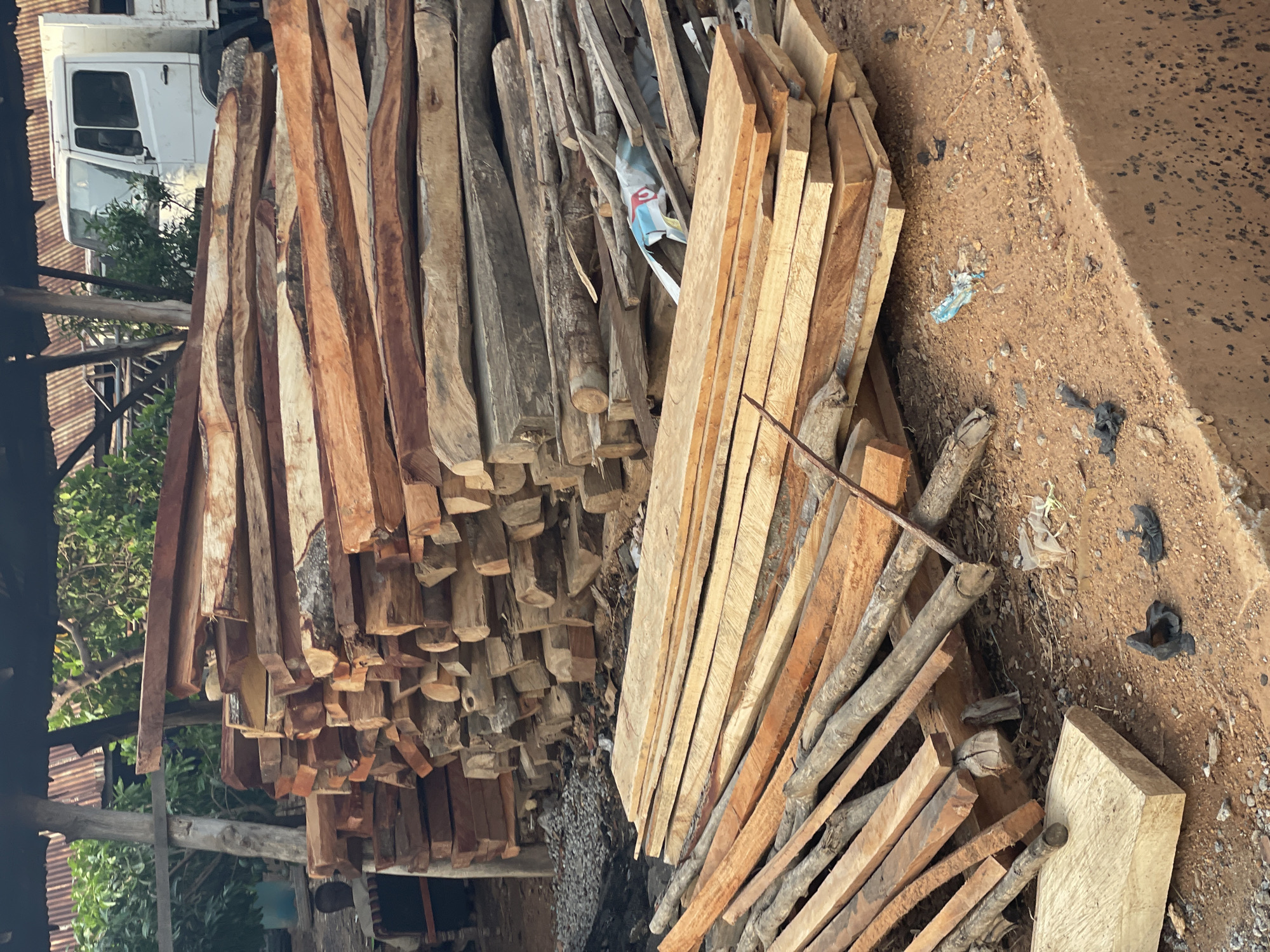 The image shows a large pile of lumber stacked on the ground in what appears to be an outdoor storage area in Akwanga, Nigeria. Two distinct piles of wood are visible: one larger pile on the left consisting of rough-cut, dark-colored lumber and another, smaller pile on the right consisting of lighter-colored, planed boards. A white vehicle is partially visible in the background, obscured by the lumber pile. The ground is covered in dirt and debris. Vegetation and a corrugated metal roof are visible in the background on the left side of the frame. There are no people visible in the image.