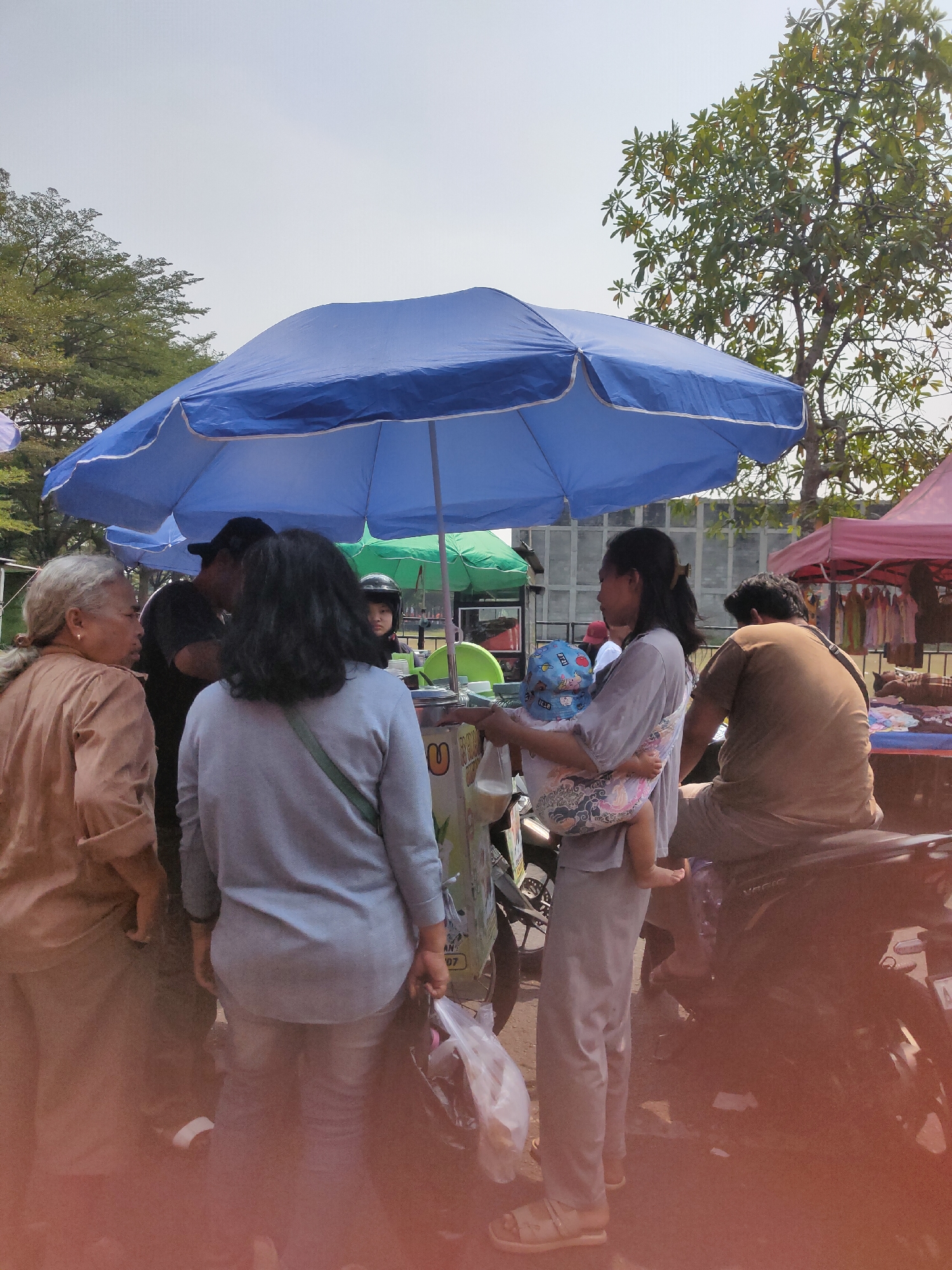 In Ngasem, Indonesia, a group of people are gathered under a large blue umbrella at an outdoor market. A woman in the foreground is holding a baby wrapped in a colorful sling, facing away from the camera. Next to her, another woman wearing a light blue shirt and jeans is holding a plastic bag. To their left, an older woman with gray hair tied back is speaking to someone obscured by others. In the background, a man in a brown shirt is seated on a motorcycle. There are other stalls with colorful tents and merchandise visible in the distance. The scene suggests a bustling market environment with active commercial transactions.