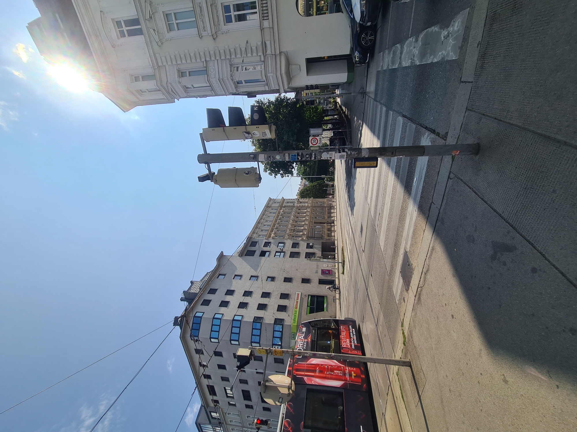 This is a street-level, eye-level, wide shot of a city intersection in Vienna, Austria, on a sunny day. The foreground shows a paved sidewalk with a prominent shadow cast by a pole. A crosswalk is visible, with tram tracks running across it. To the left, a red tram with advertisements for L'Oréal is passing by. In the background, there are several buildings of varying architectural styles, some with modern designs and others with more classical features. The sky is clear blue with some wispy clouds and the sun is shining brightly, causing lens flare. There are traffic lights and street signs present. A few trees line the street in the distance. There are no people in focus.