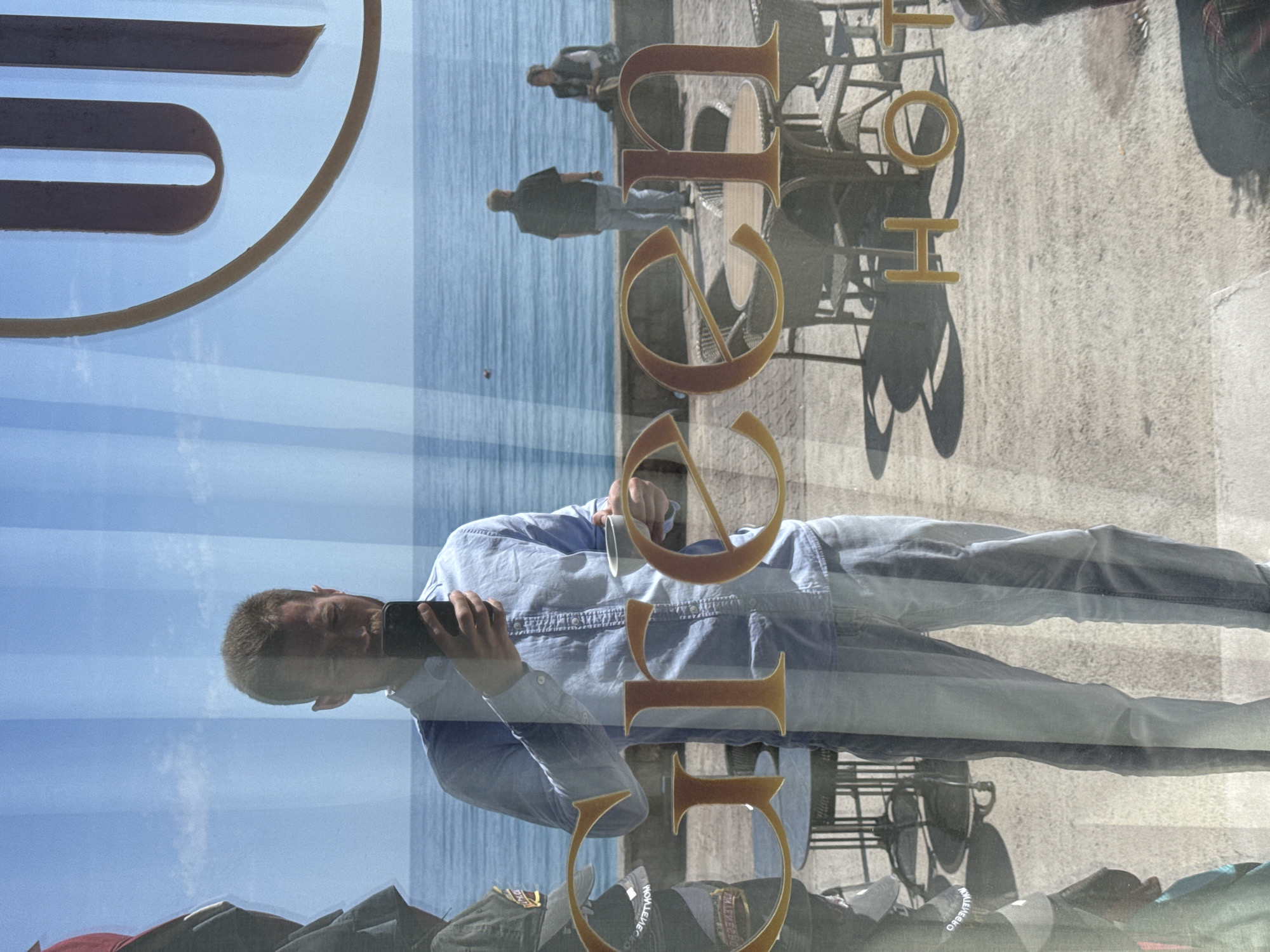 The image captures a complex, multi-layered reflection on a glass surface, likely a shop or restaurant window, presented with a 90-degree clockwise rotation. In the foreground, the reflection of a man (the photographer) is prominent. He is seen from the chest up, holding a smartphone as if taking a selfie or photographing the scene. He is wearing a light blue collared shirt and blue jeans, with a slight smile on his face.
Decorative golden letters are affixed directly to the glass. The word "HOTEL" is clearly visible, oriented vertically, running down the right side of the frame. Below "HOTEL," a large "G" is discernible, followed by partial letters that appear to be part of a word like "GALEB" or similar. Other fragmented golden letter shapes are present in the top-left section.
Reflected in the background is a bright outdoor scene typical of a coastal area, specifically Rafailovići, Montenegro. A calm, blue sea occupies the upper portion of the reflected view (appearing horizontally if the image were rotated counter-clockwise to its natural orientation). Below the sea, a sunny promenade or sidewalk is visible, populated by several reflected figures, including two individuals walking away from the viewer. Wicker-style outdoor tables and chairs are also reflected, casting sharp, distinct shadows on the light-colored pavement, indicating strong sunlight and clear weather. The sky above the sea is a clear blue with subtle white clouds.
The image is a dynamic composition showcasing the interplay of the photographer's reflection, the fixed text on the glass, and the vibrant external environment.