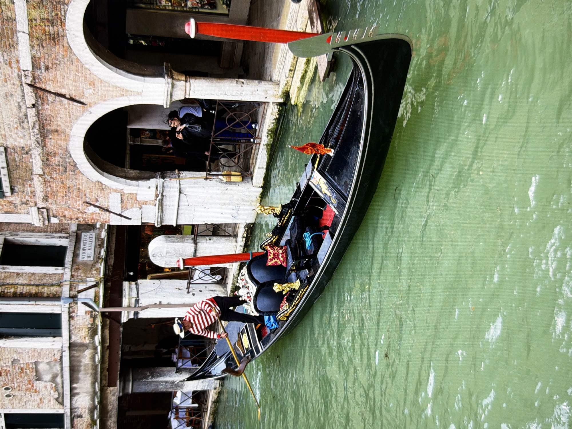 This image depicts a gondola being rowed on a canal, characteristic of Venice, Italy, not Kyiv, Ukraine. A gondolier in a red and white striped shirt and a straw hat stands at the stern, propelling the gondola with a single oar. The gondola itself is black with ornate gold detailing and red upholstery on its seats. In the background, a building with brick facade, arched walkways, and balconies overlooks the canal. Some individuals are visible standing on a balcony, with one person taking a photograph. Tables and chairs are set up on another side, suggesting a restaurant or cafe. The water of the canal is a greenish hue, reflecting the surrounding architecture.