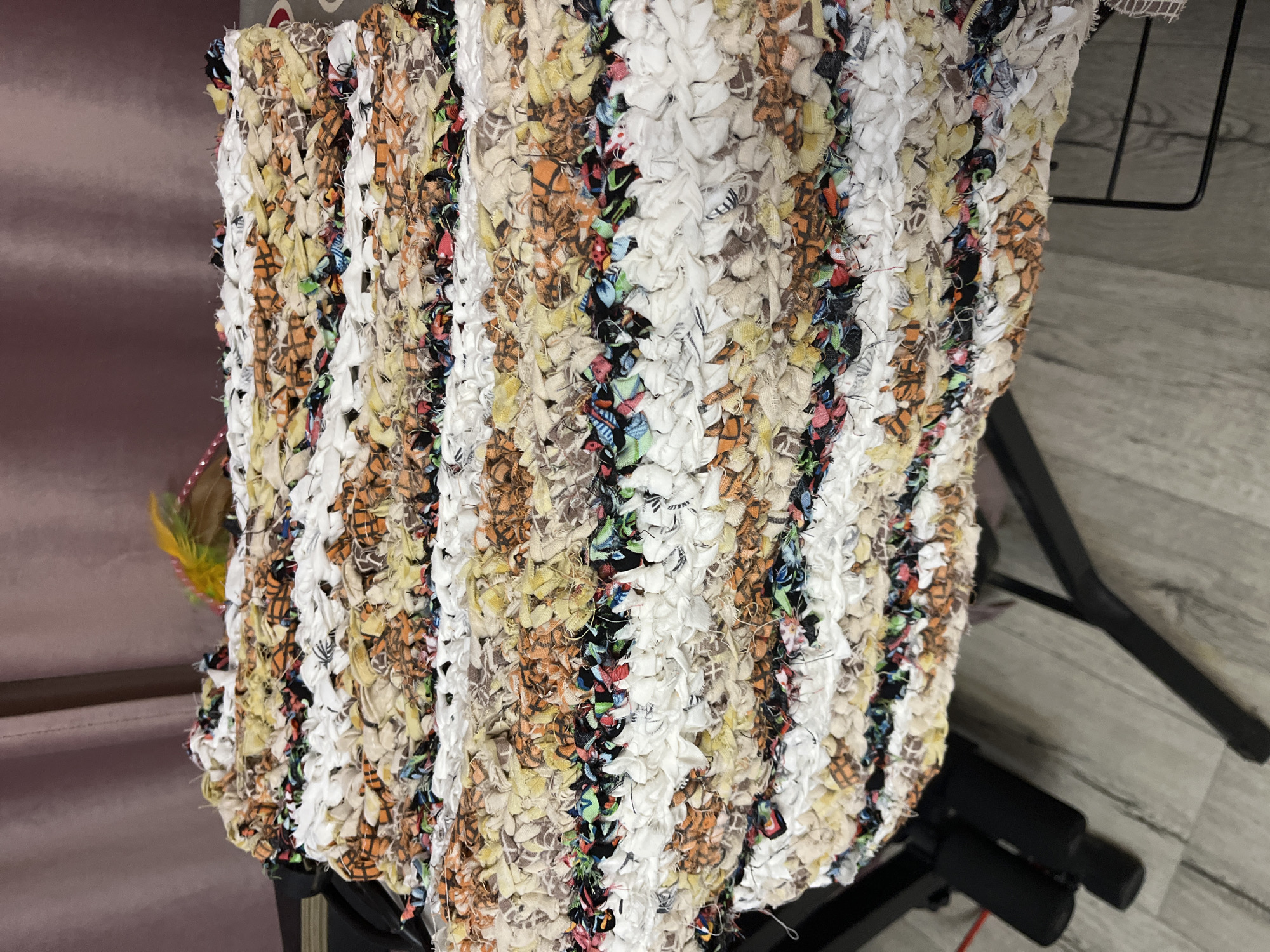 The image shows a close-up, high-angle view of a handmade rag rug. The rug is made of woven strips of fabric in various colors and patterns, including white, beige, orange, and black. The texture is rough and uneven due to the nature of the fabric strips. A small yellow and green feather is visible on the top of the rug. The rug is placed on a dark, metallic stand, which is partially visible in the lower part of the image. The background consists of a muted pink curtain and a light-colored wooden floor. There are no people visible in the image. The scene appears to be indoors, likely in a domestic setting. The specific location within Krasnyye Barrikady, Russia cannot be determined from the visual information provided.