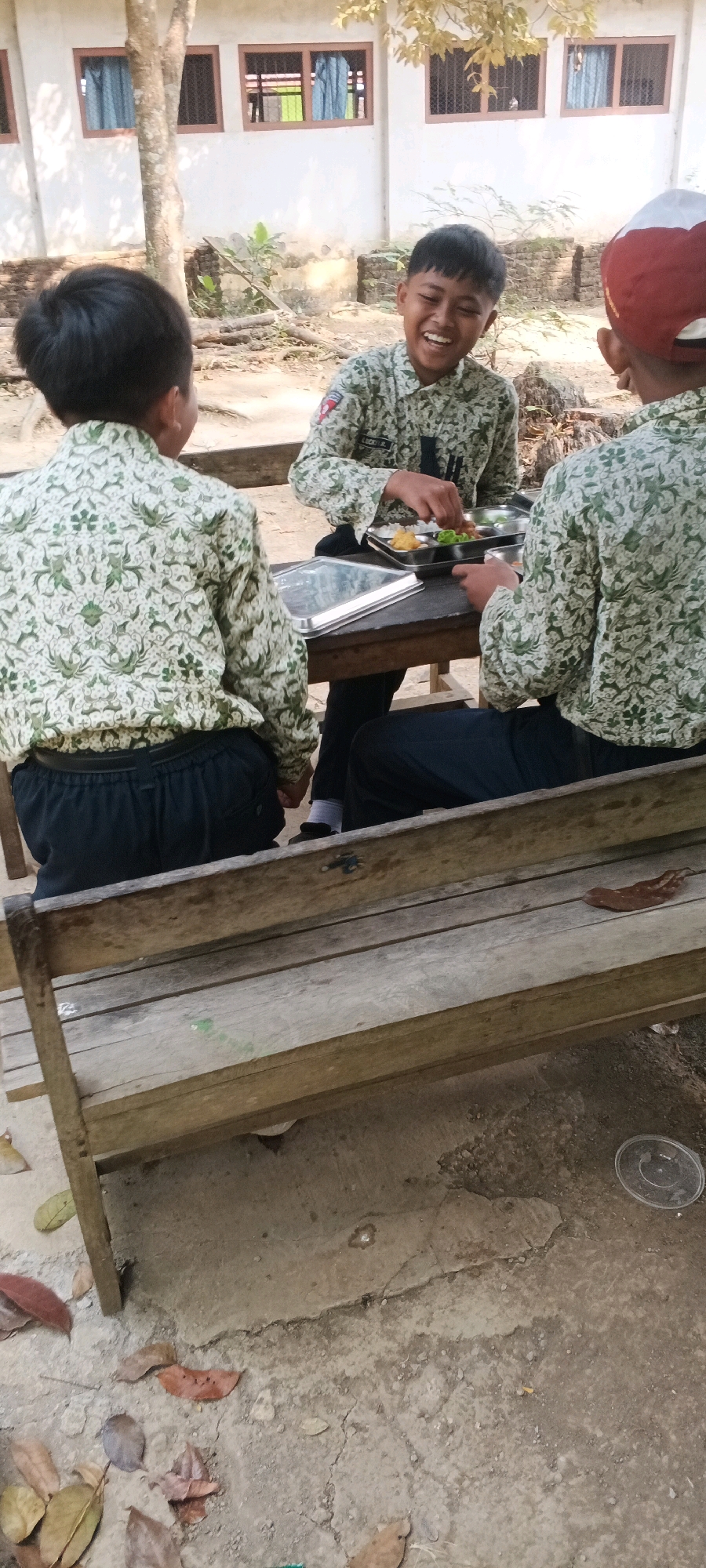 Three young boys in school uniforms are sitting at a wooden table outside a building. The boy in the center is laughing heartily, with his mouth wide open. He is wearing a light-colored shirt with a green leaf pattern and dark pants. His hands are over a metal tray filled with food. To his left, another boy, seen from behind, is wearing a similar uniform and dark pants. To his right, a third boy, also seen from behind, wears the same patterned shirt and dark pants, and a red and white cap. The background shows a white building with windows and a tree. The ground is rough, a mix of dirt and concrete, with fallen leaves scattered around. A wooden bench is in the foreground. The scene appears to be in the city of Penanggungan, Indonesia.