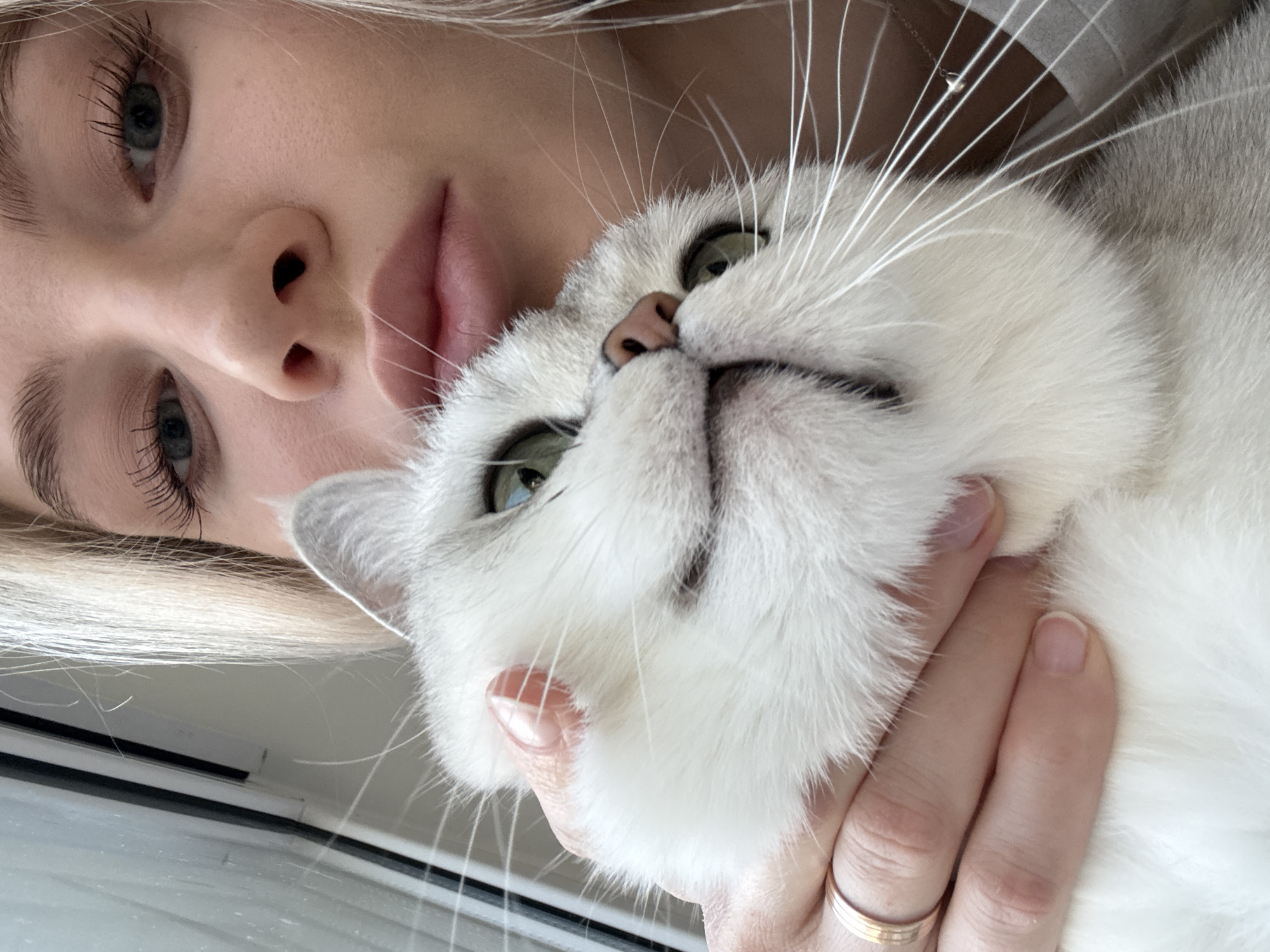 A young woman with fair skin, blonde hair, and heavily mascaraed eyelashes is holding a white and gray cat close to her face. Her lips are slightly parted, and her nose is close to the cat's fur. The cat has green eyes and a pink nose. The woman is wearing a beige or gray top, and a thin gold necklace is visible. She's also wearing a gold ring. The background includes a window frame, and it is likely an indoor setting. There is no readily available information to indicate that this picture was taken in Izhevsk, Russia.