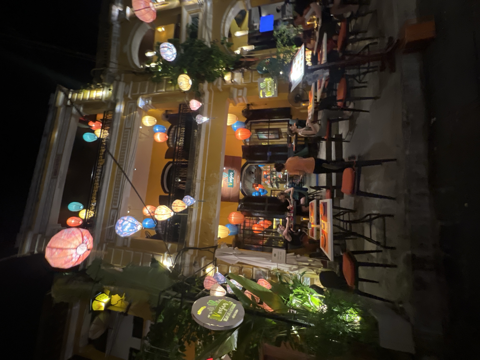 The image depicts a nighttime scene at a vegan cafe and restaurant in Hoi An, Vietnam. The building is painted a vibrant yellow and is illuminated by numerous colorful lanterns, including red, pink, blue, and yellow, hanging from the balconies and surrounding the entrance. Tables and chairs are set up outside the establishment, some occupied by patrons. A sign with the restaurant's name, "Vegan," is visible on the left side of the image, adorned with a green leaf logo. The street is paved and dark, with the lighting primarily coming from the restaurant and its lanterns. Plants are strategically placed around the outdoor seating area, adding to the ambiance. In the foreground, tables are set with placemats and what appear to be candles. People are seen dining and socializing at the tables. The overall atmosphere suggests a lively evening dining experience in Hoi An.