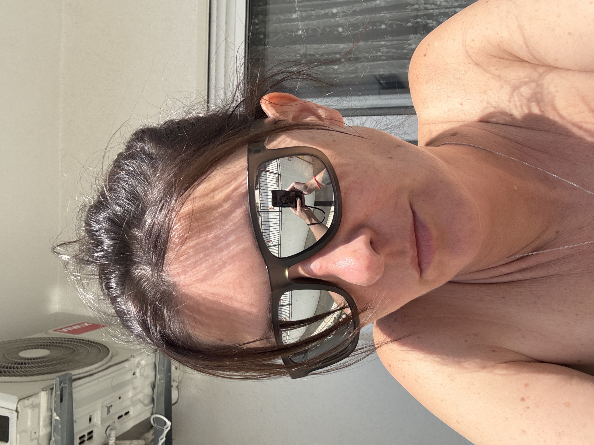 The image shows a selfie of a light-skinned person with dark hair, wearing sunglasses. The person's bare shoulder and part of their chest are visible. The reflection in the sunglasses reveals a figure holding a phone, presumably taking the picture, in an outdoor setting in Bar, Montenegro, as seen by the buildings and architecture. A window with blinds is behind the person, and an air conditioning unit is partially visible in the lower-left corner.
